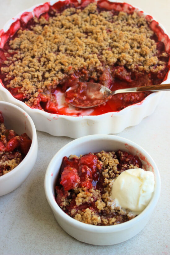 White bowl with strawberry crisp and a scoop of ice cream. Behind, a round baking dish with strawberry crisp.