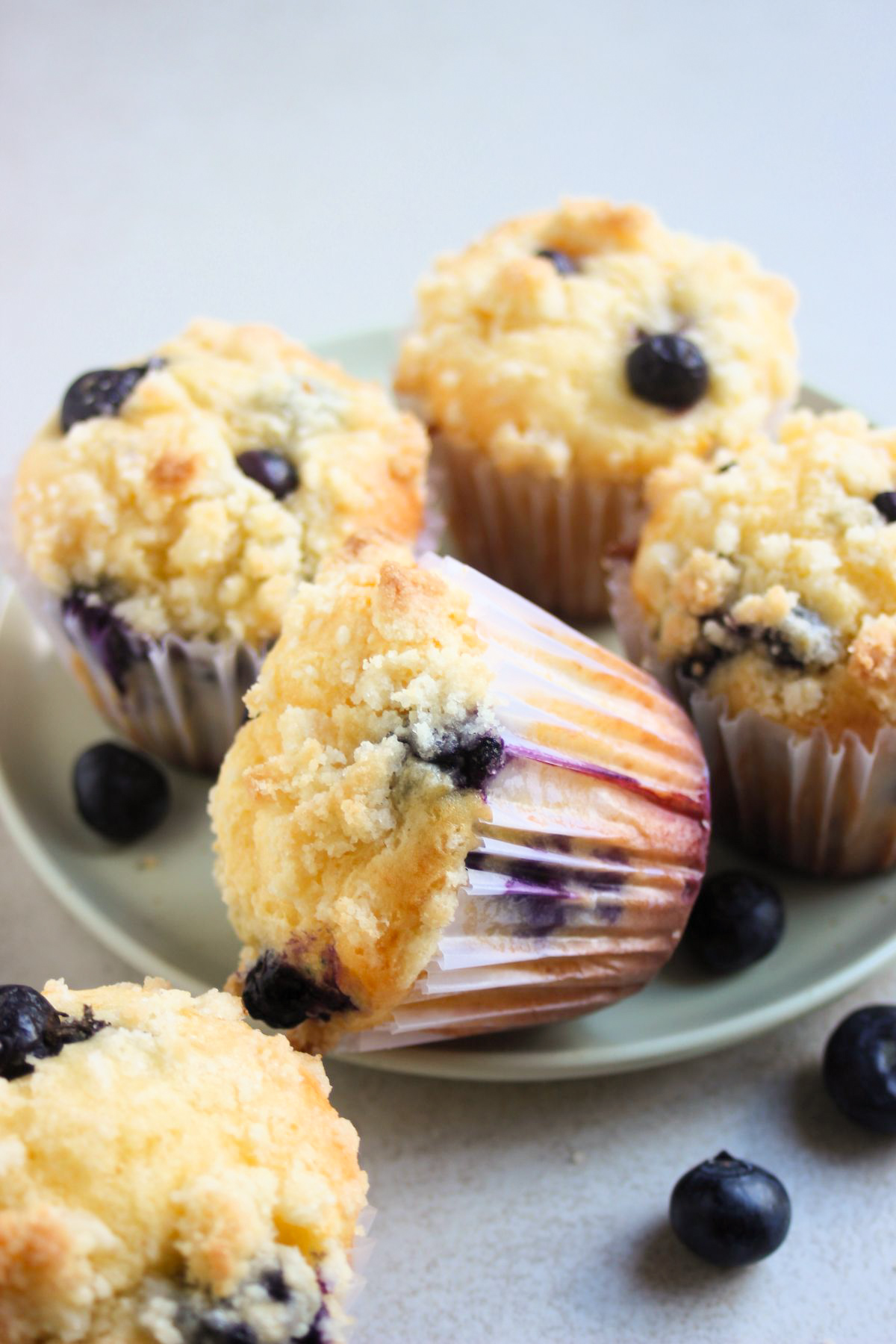 Blueberry streusel muffins in liners on an aqua-green plate. One is lying down. Fresh blueberries scatter around.