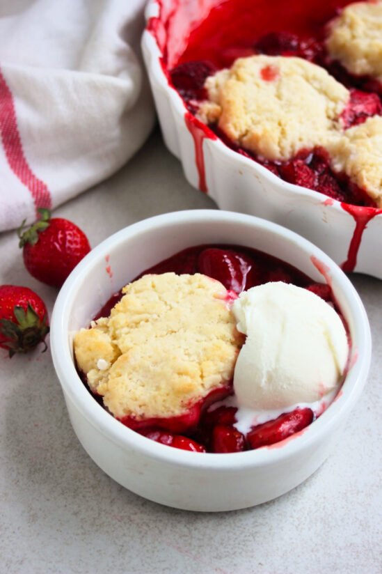Small bowl with strawberry cobbler and a scoop of ice cream. Baking dish with cobbler beside, a kitchen towel, and fresh strawberries.