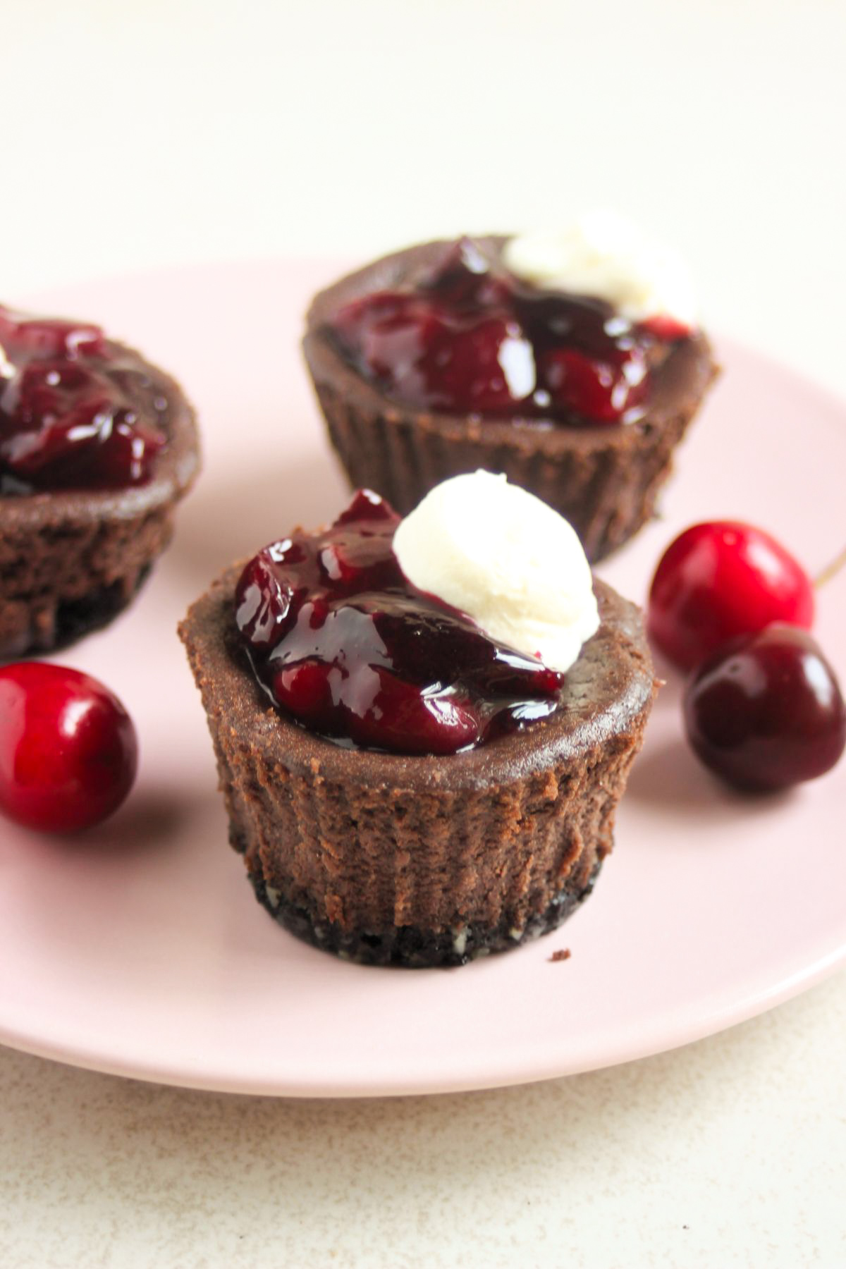 Mini black forest cheesecakes with cherry topping and whipped cream on a pink plate. Fresh cherries on the sides.