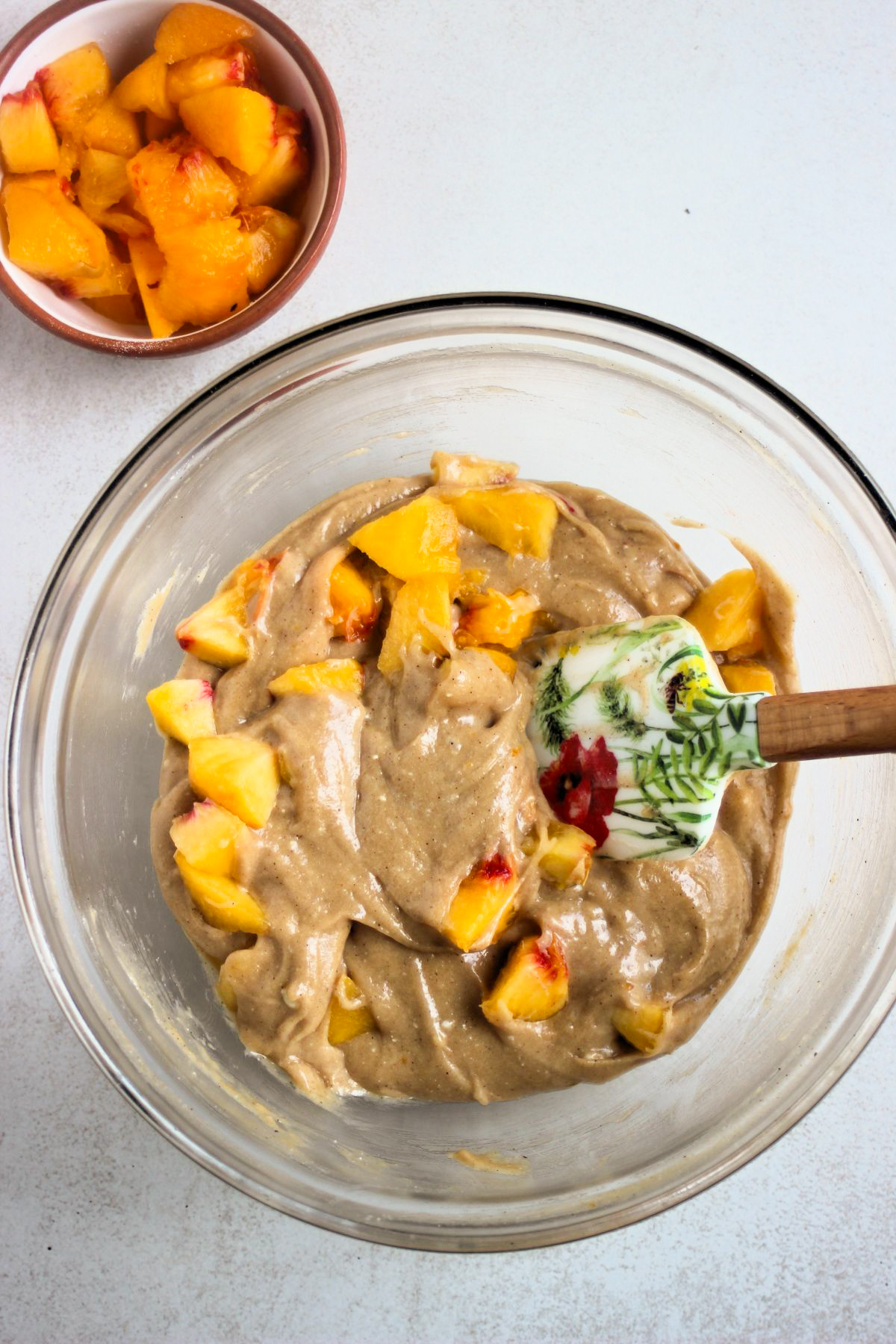 Glass bowl with a mixture of sliced peaches and a light brown batter. A rubber spatula.