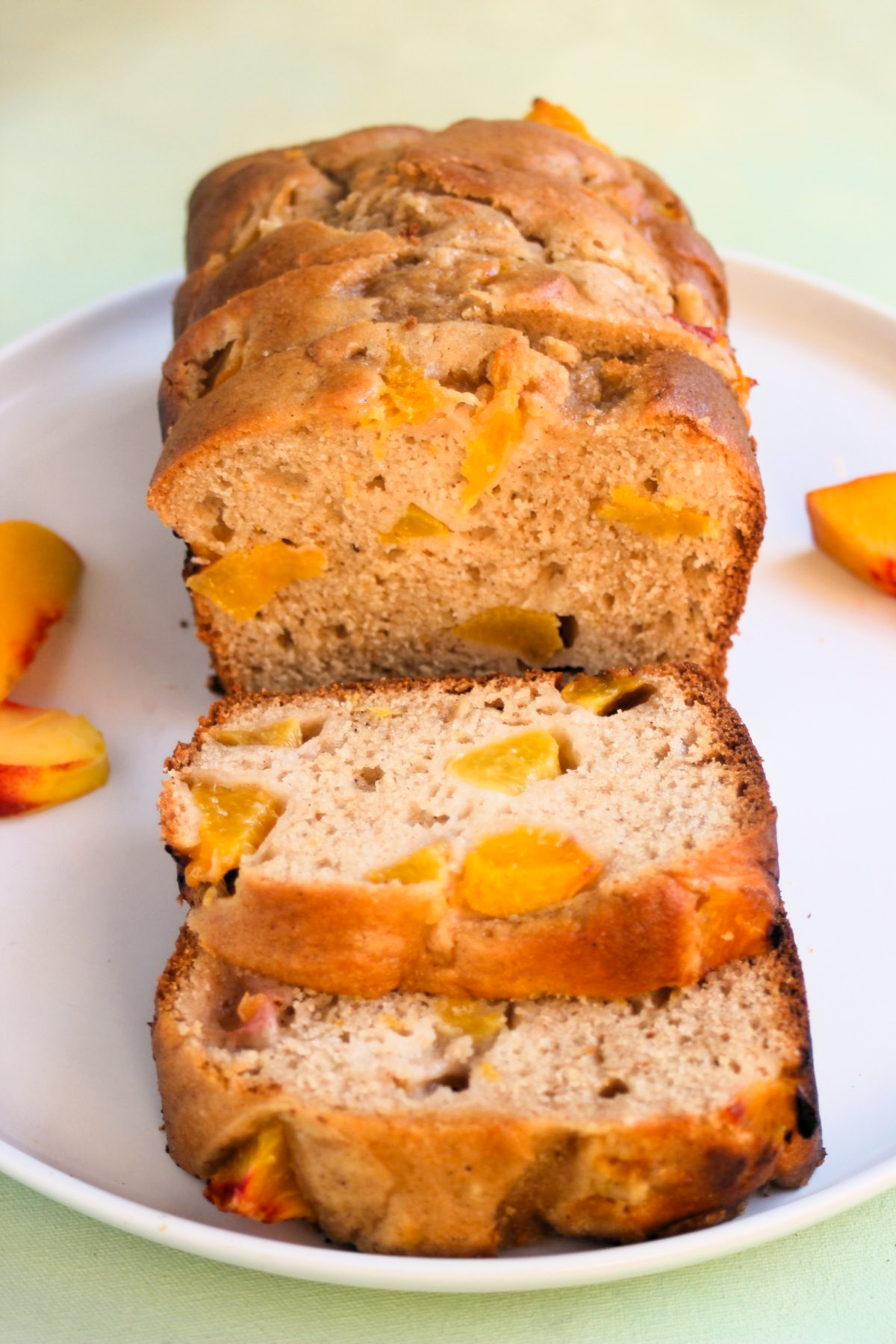 Peach loaf cake and slices of it, on a white plate. Fresh peach slices on the sides.