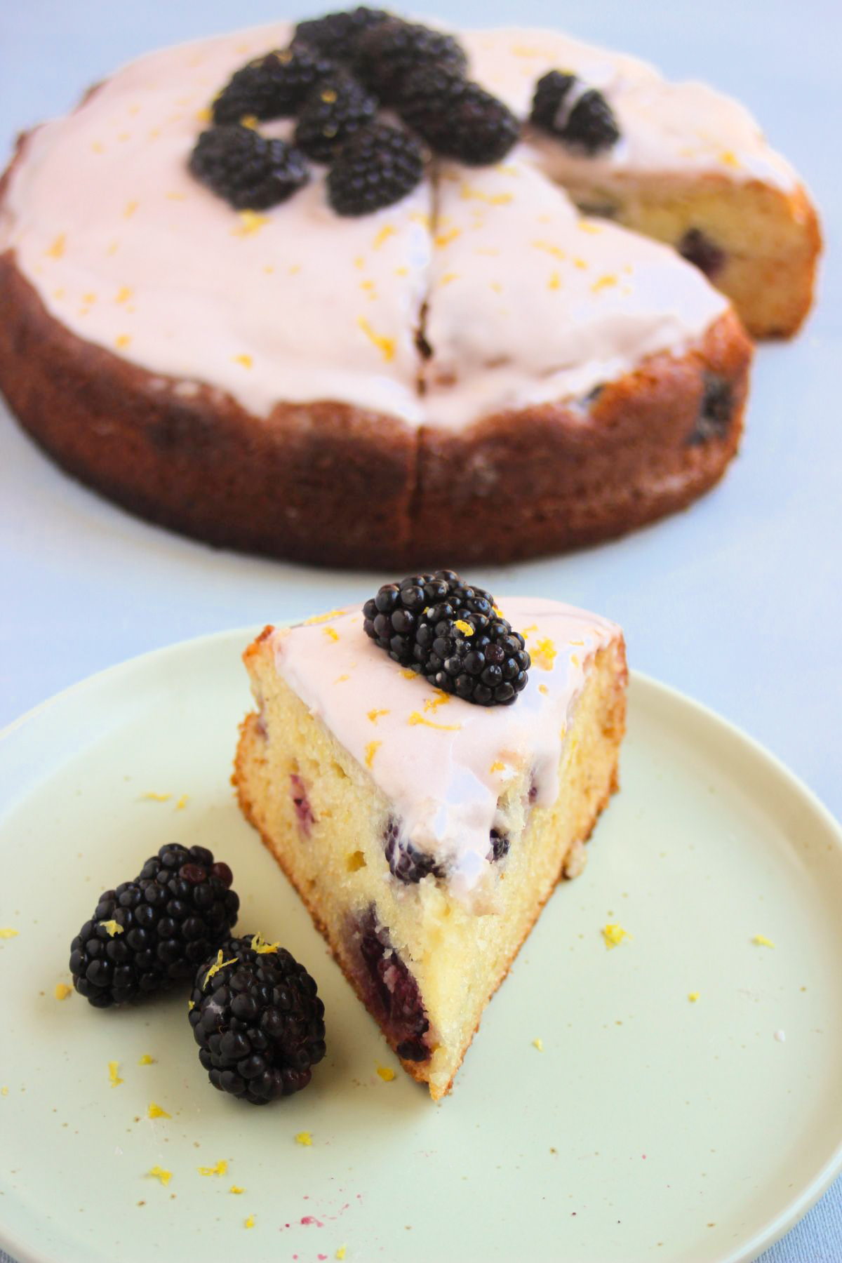 Portion of blackberry cake with frosting and fresh blackberries on an aqua-green plate. The cake is behind.