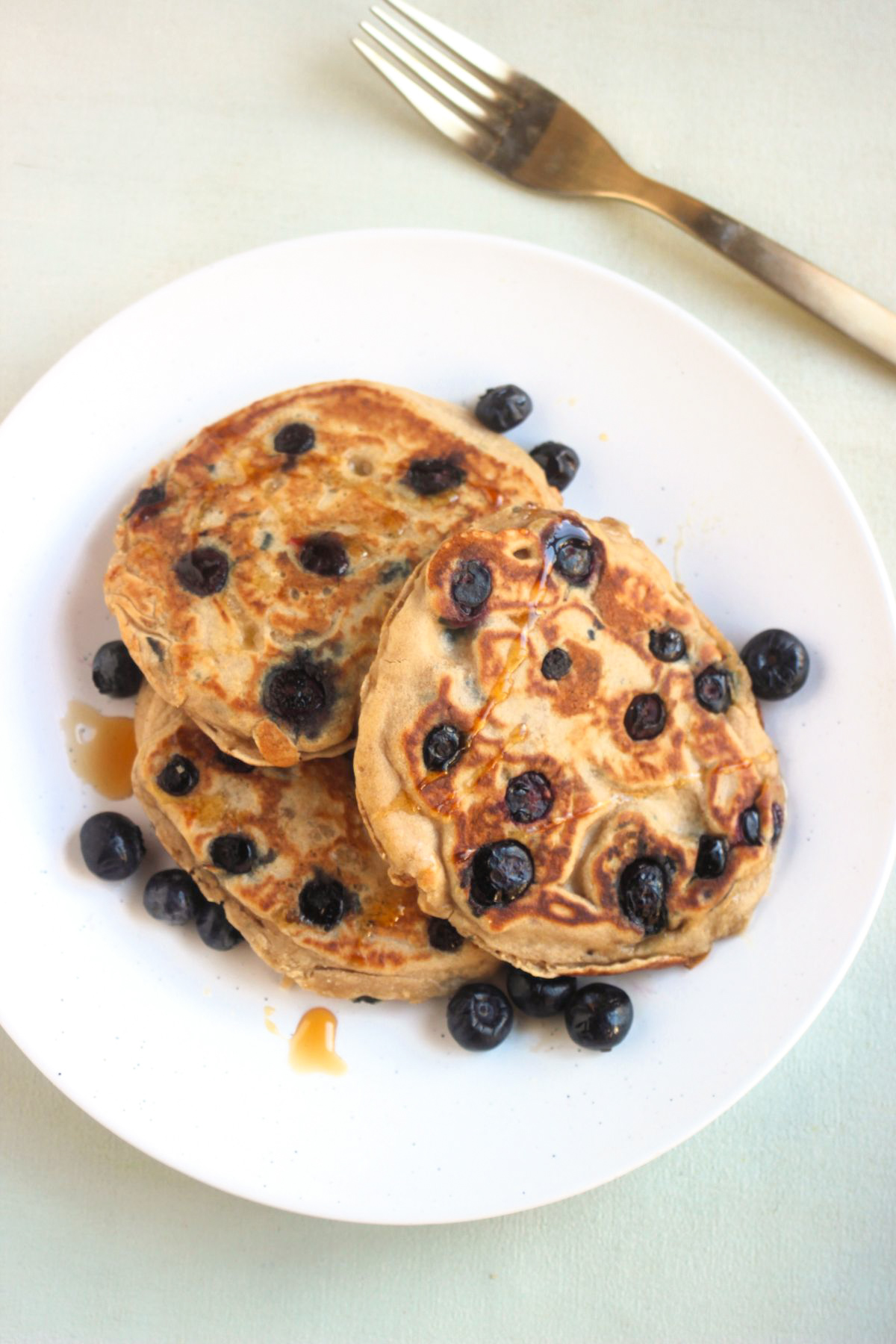 Top view of three blueberry pancakes on a white plate and scattered blueberries. Golden fork next to the plate.