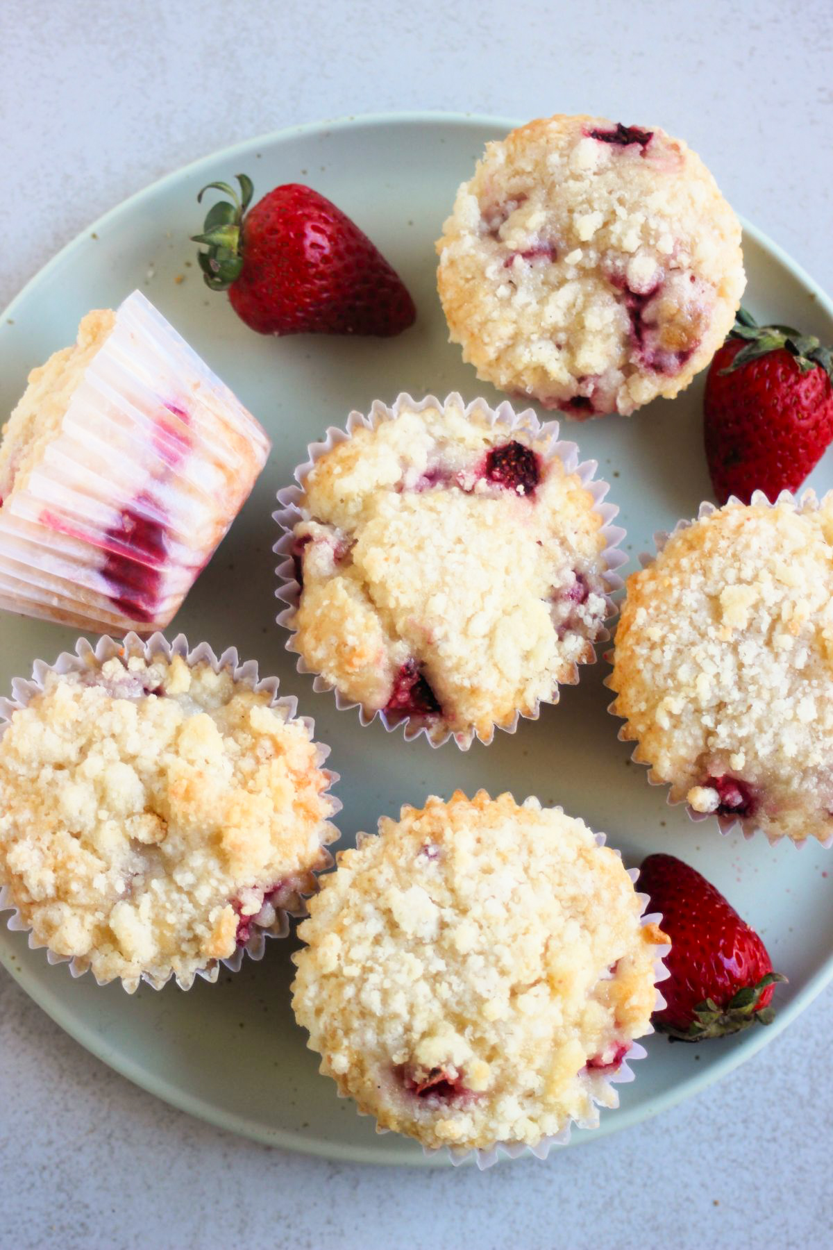 Strawberry streusel muffins with liners on an aqua green plate seen from above. Fresh strawberries on the sides.