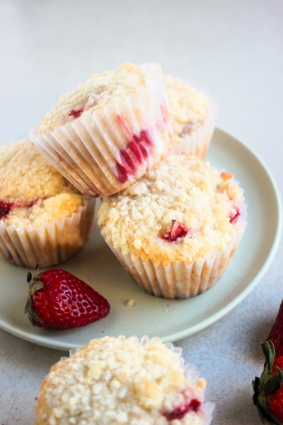 Strawberry streusel muffins with liners on an aqua green plate. One muffin is above the others. A fresh strawberry.