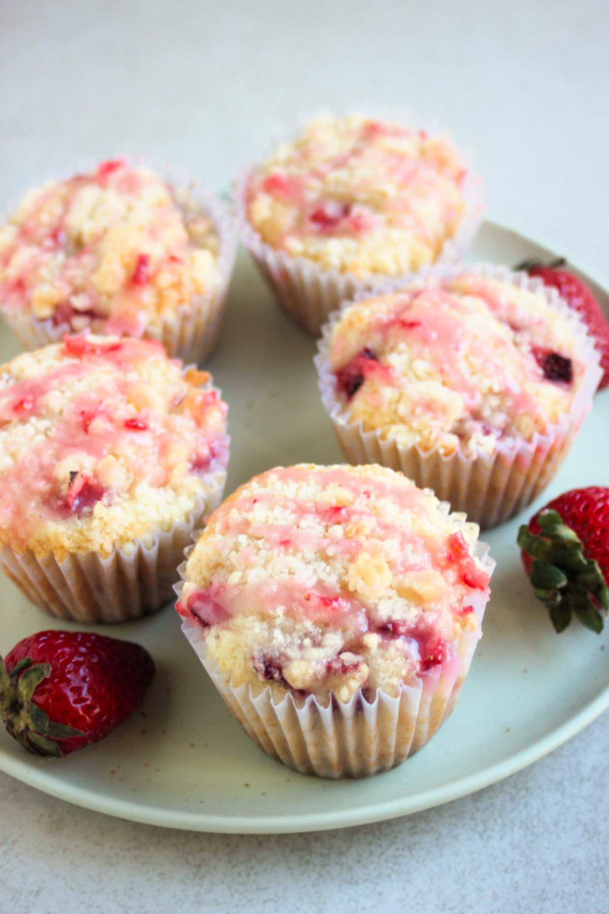 Strawberry streusel muffins with liners and strawberry icing on an aqua green plate. Fresh strawberries on the sides.