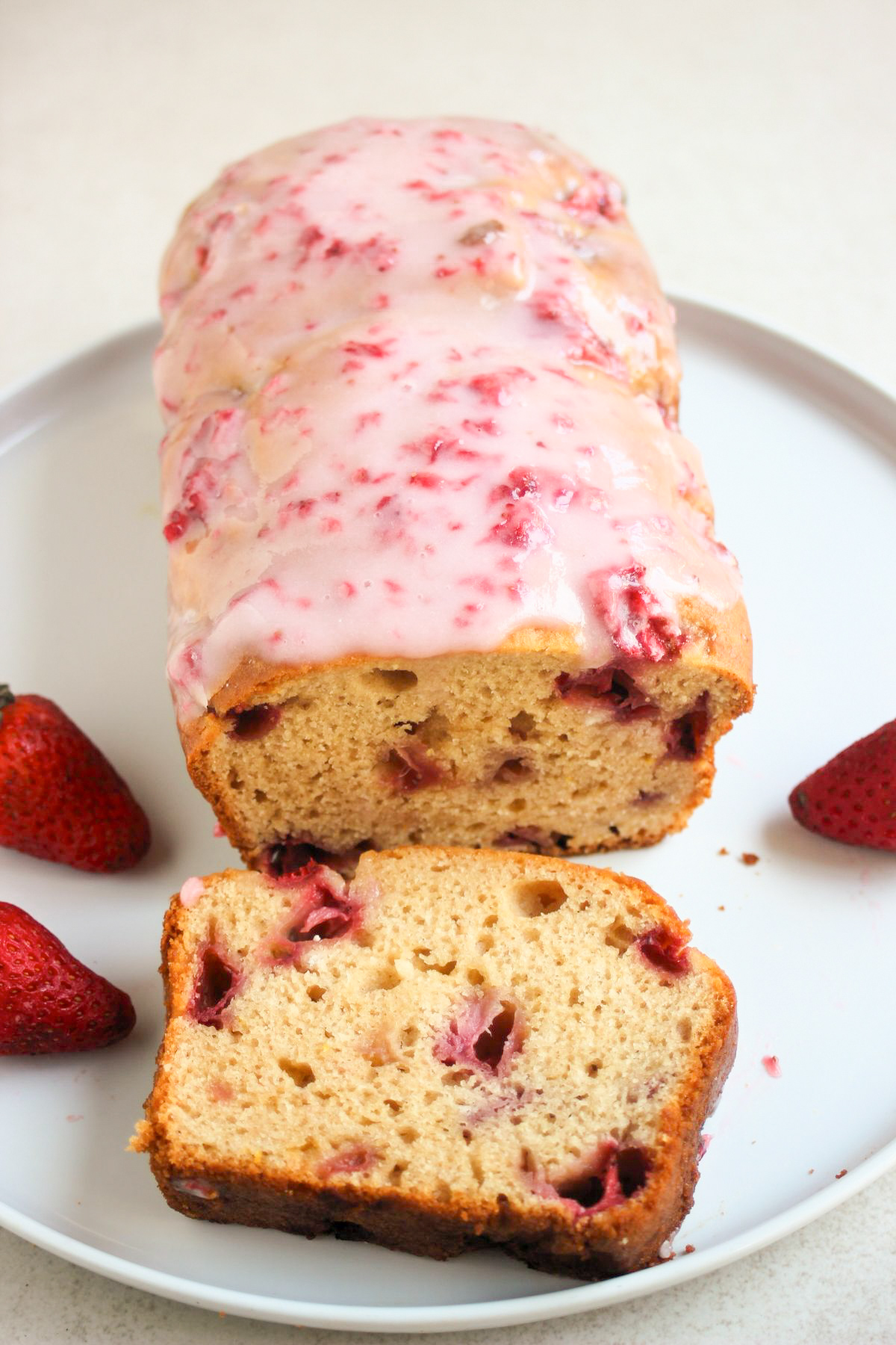 Strawberry bread with pink icing, and a portion of it on a white plate. Fresh strawberries on the sides.