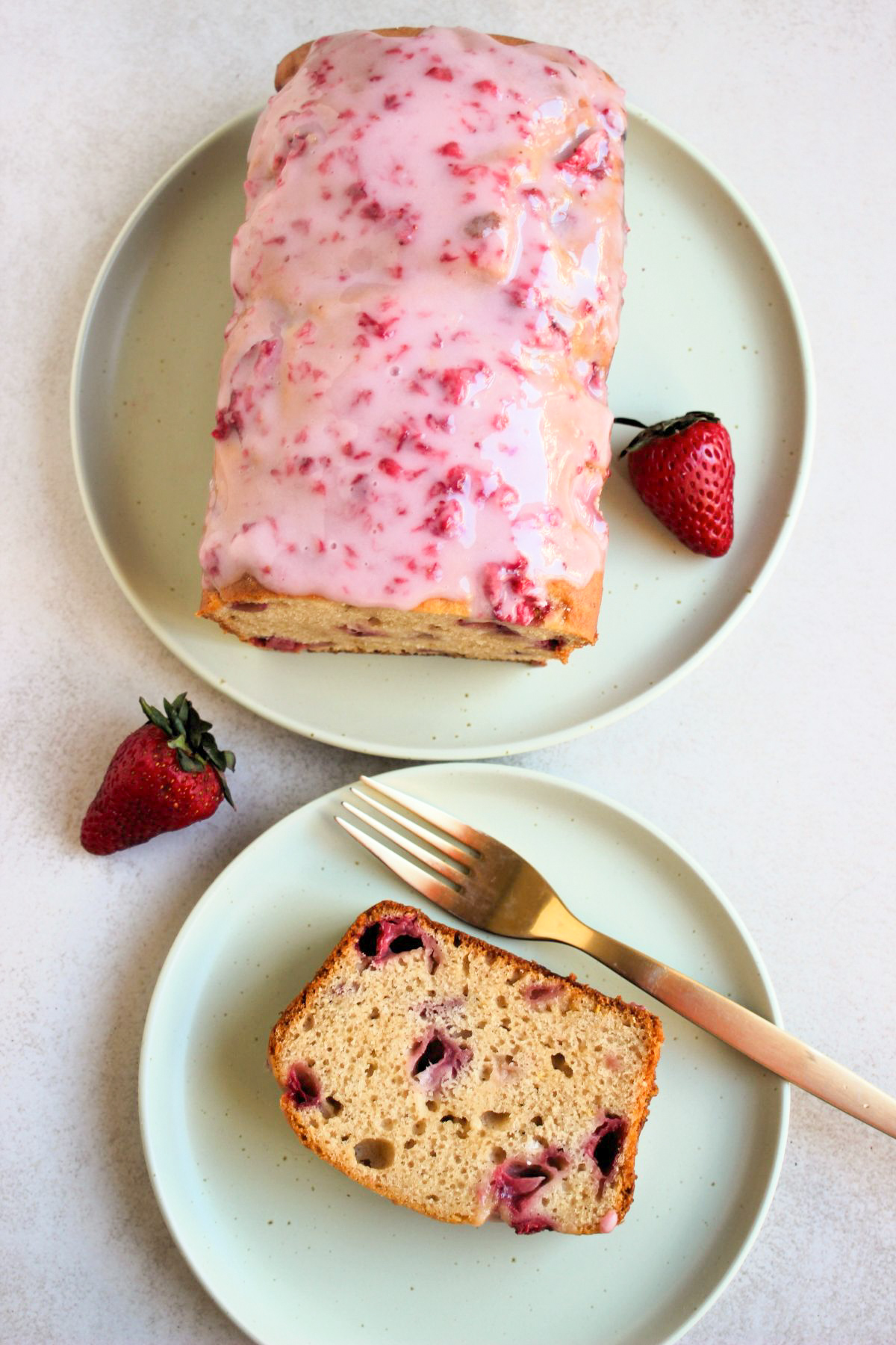 Top view of strawberry bread with pink icing on an aqua green plate, and a portion of it and a golden fork on a small aqua-green plate.