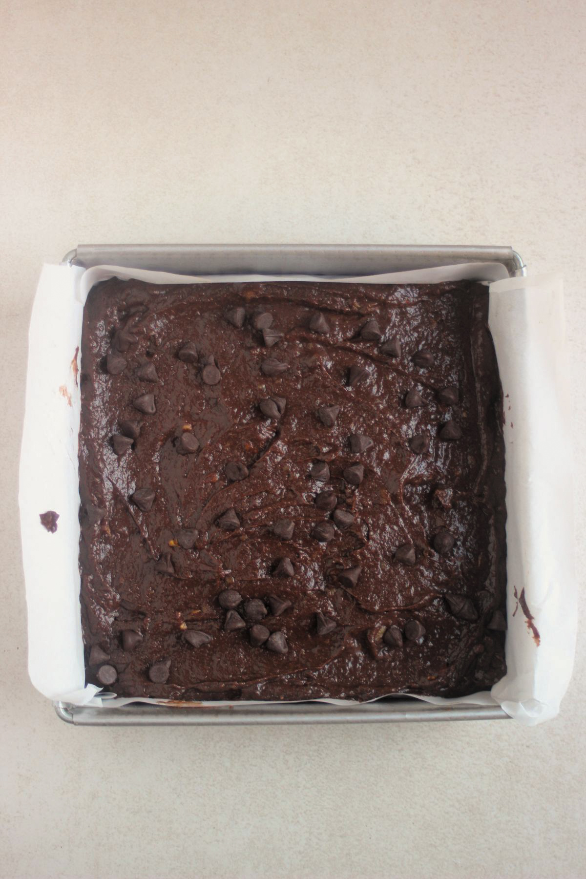 Overhead view of a square dark chocolate brownie in a parchment paper in a square pan, topped with scattered chocolate chips.