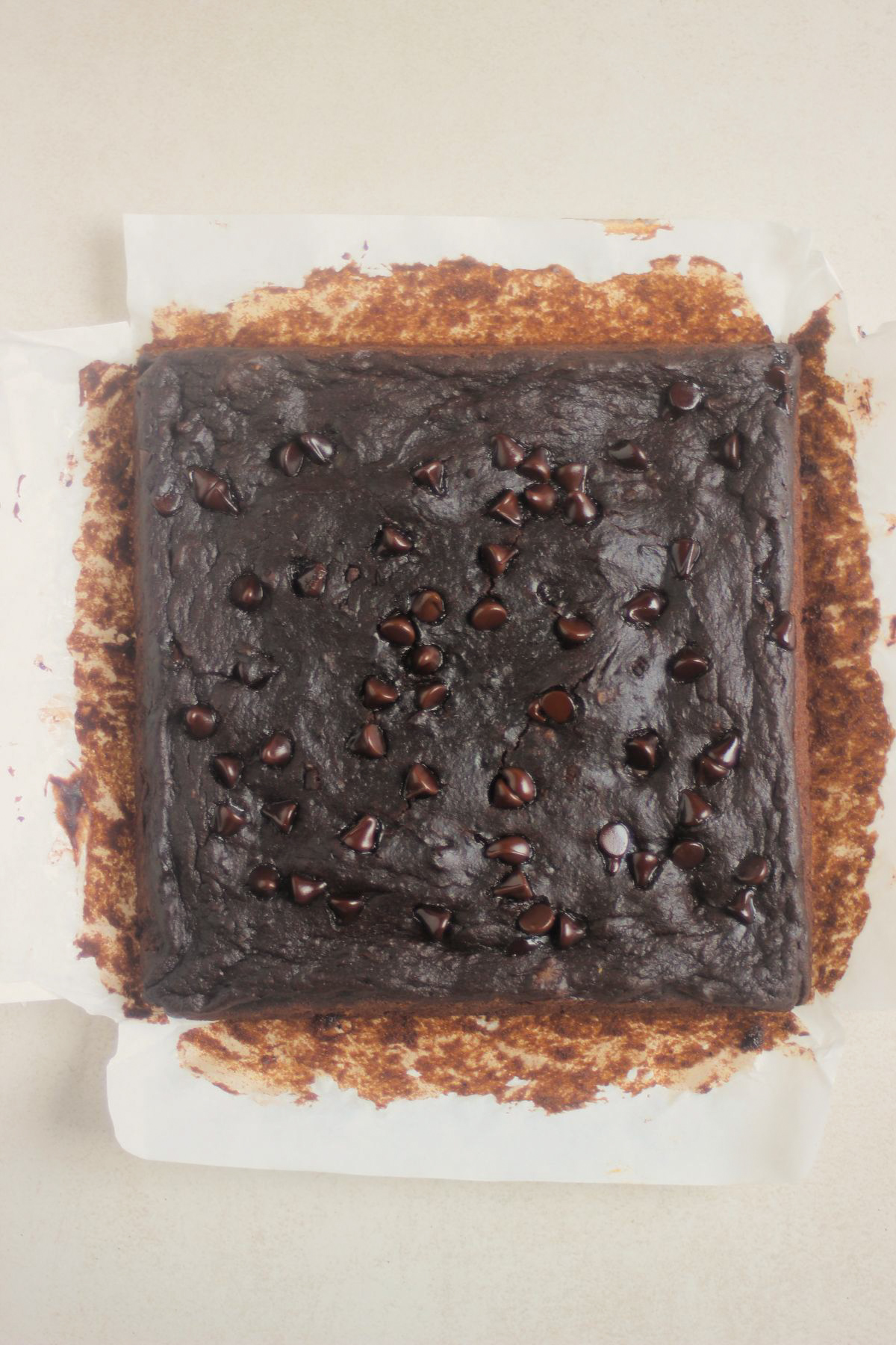 Overhead view of a square dark chocolate brownie in parchment paper, topped with scattered chocolate chips.