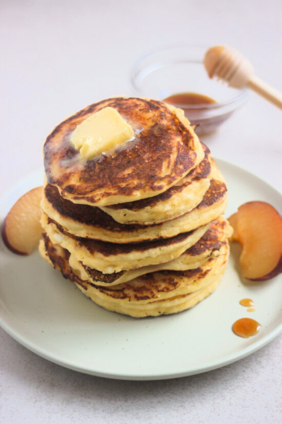 Stack of cottage cheese pancakes with a cube of butter on top, and fresh plum slices on the sides on a white plate.