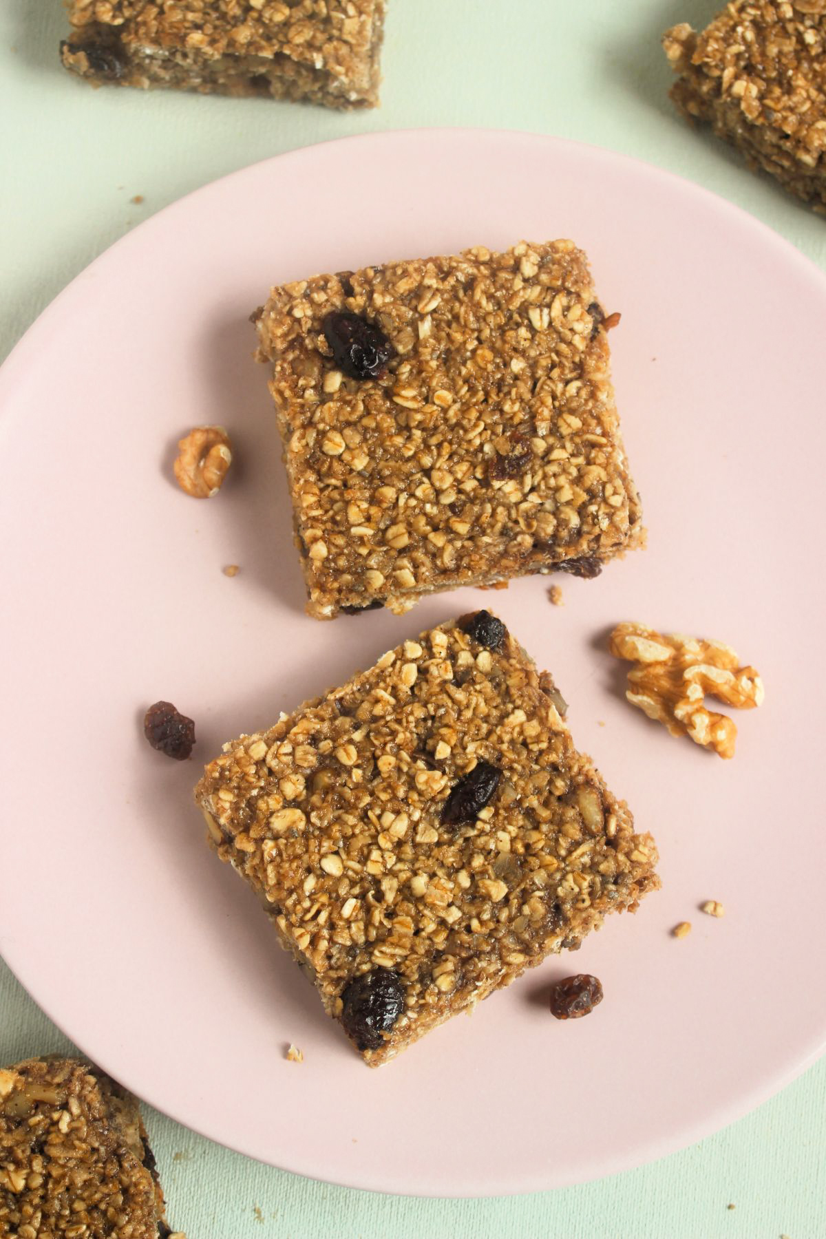 Top view of two baked oatmeal bars on a pink plate. Raisins and walnuts are scattered around.