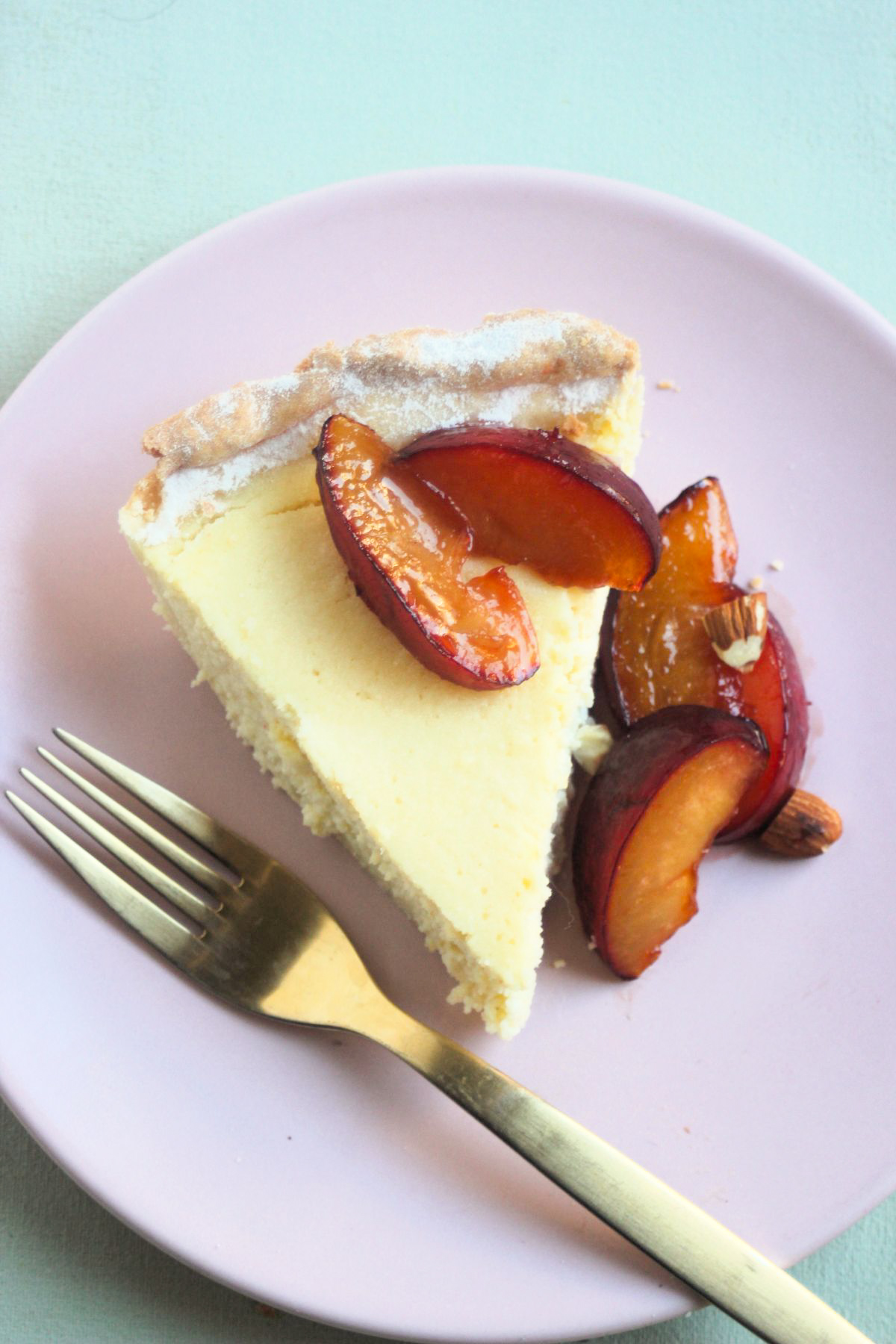 Top view of a ricotta pie portion on a pink plate with plums and a golden fork.