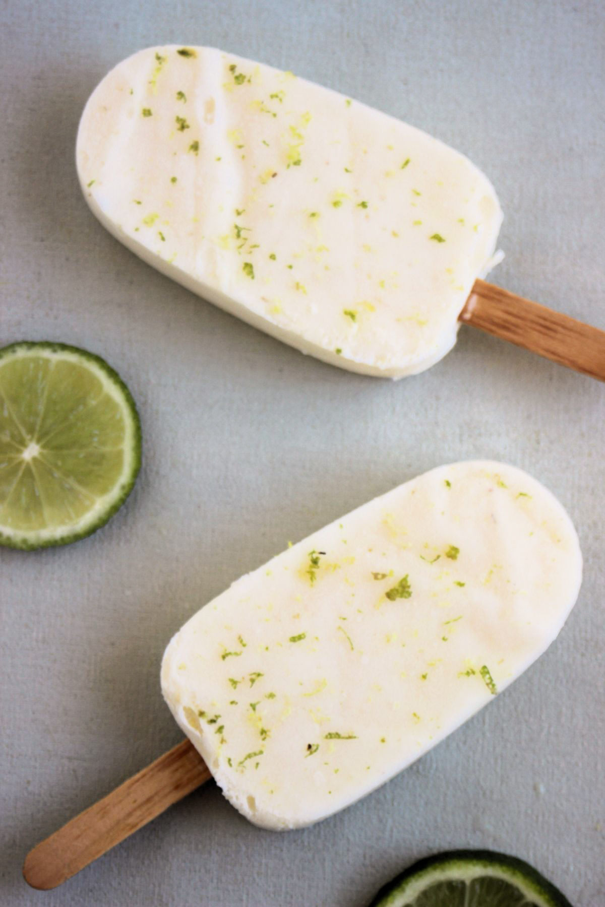 Top view of two lime popsicles on a light-grey surface and two fresh lime slices.