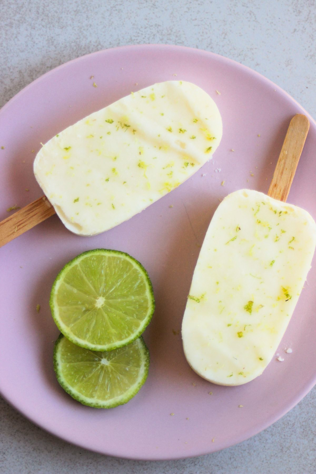 Top view of two lime popsicles on a pink plate, with two fresh lime slices.