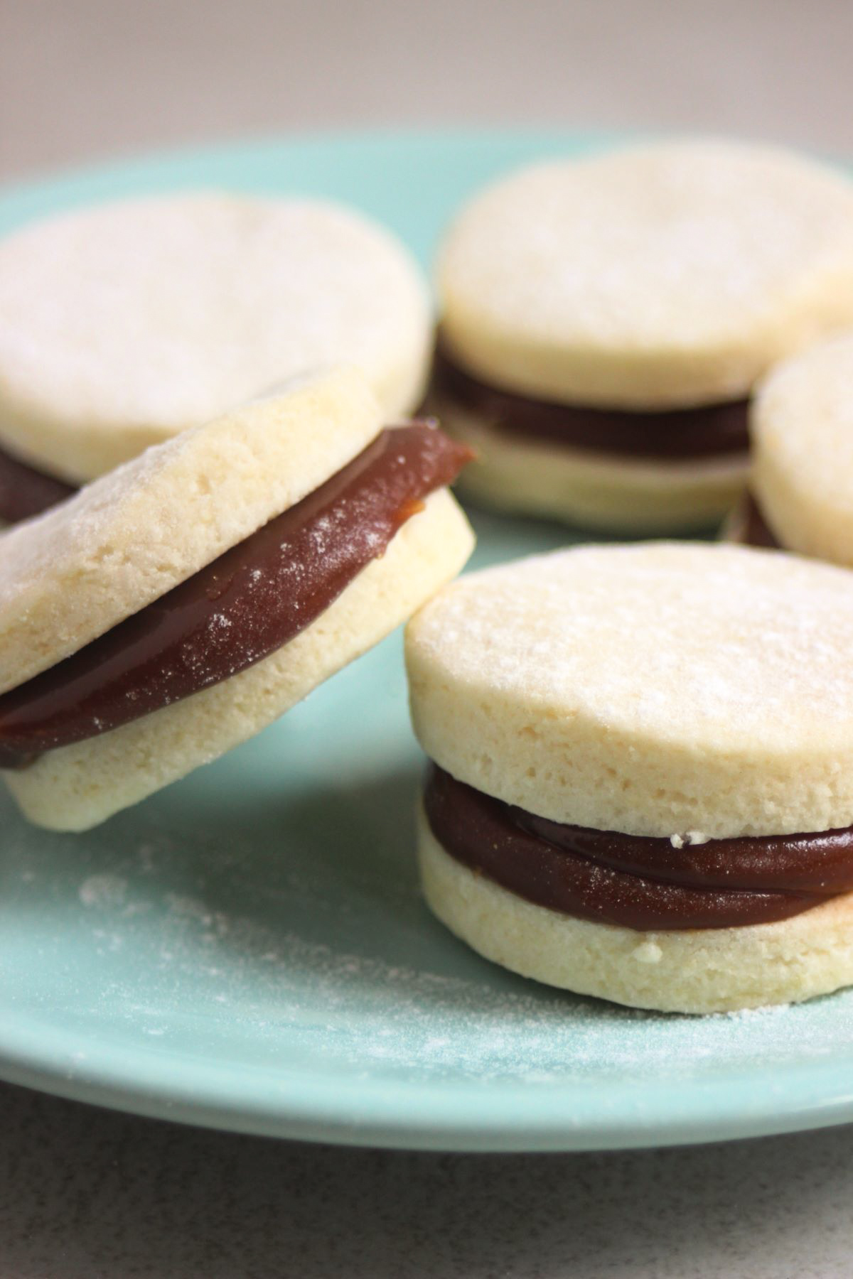 Almond alfajores on a green aqua plate. One alfajor is resting on another alfajor.