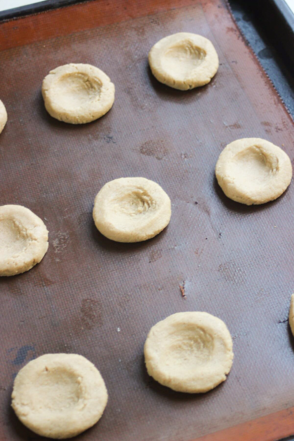Slightly flattened scoops of cookie dough with an indentation in the middle of the cookie on a silicone mat.
