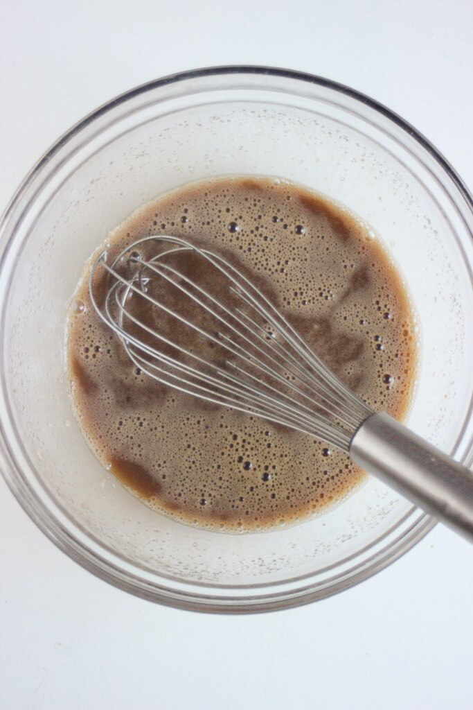 Glass bowl with a brown liquid and a hand whisk.