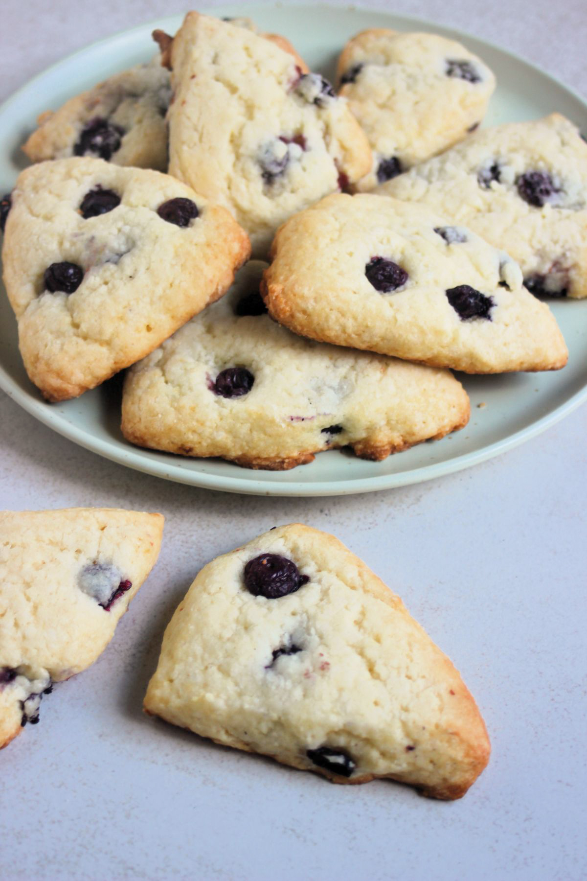 Blueberry scon on a white surface more scones behind on an aqua-green plate.