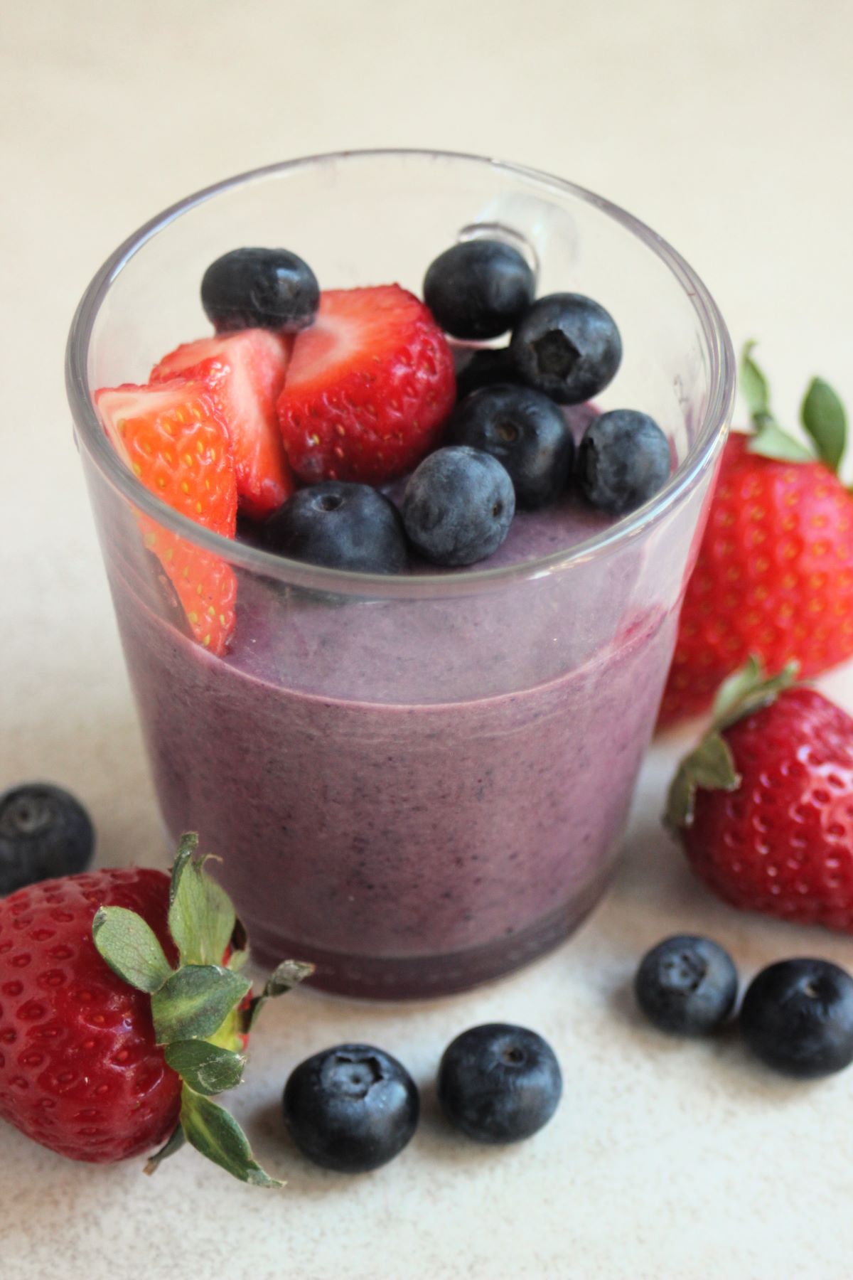 Glass cup with blueberry panna cotta, blueberries, and strawberries. More strawberries and blueberries are scattered around.