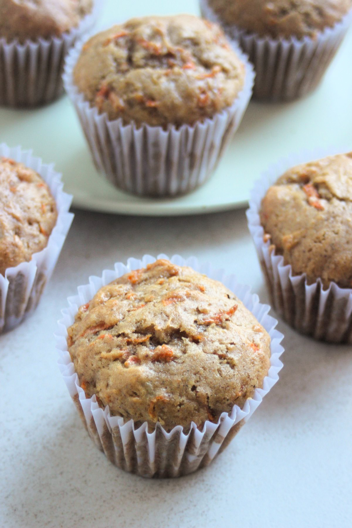 Carrot cake muffins in liners on a white surface. More muffins behin on an aqua-green plate.