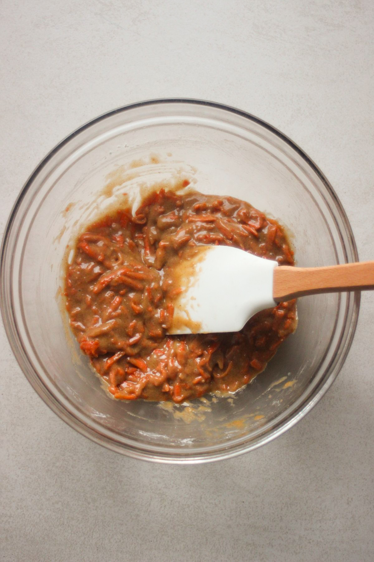 Glass bowl with a mixture that includes grated carrtos and a rubber spatula.