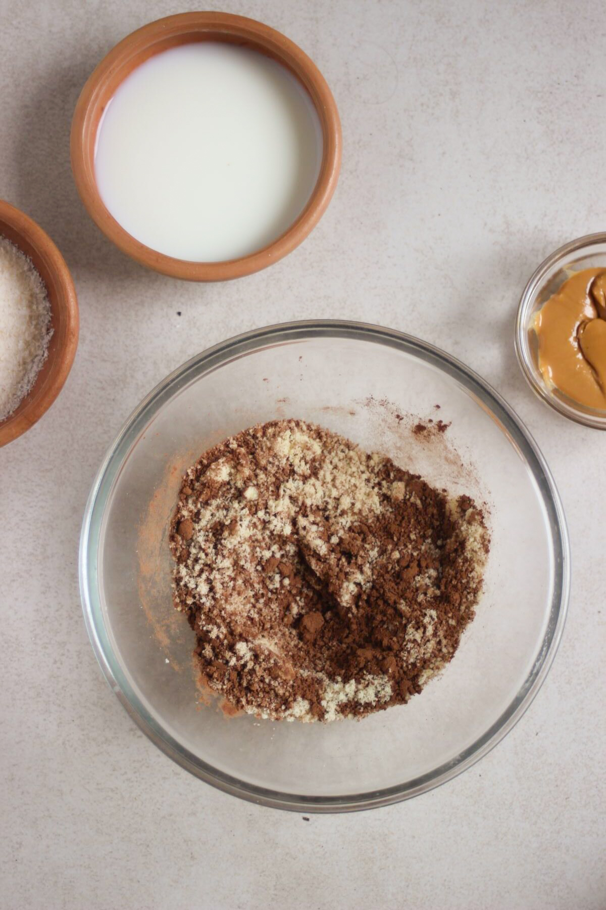 Glass bowl with cocoa powder, almond flour, and shredded coconut. Another pot with white liquid beside.