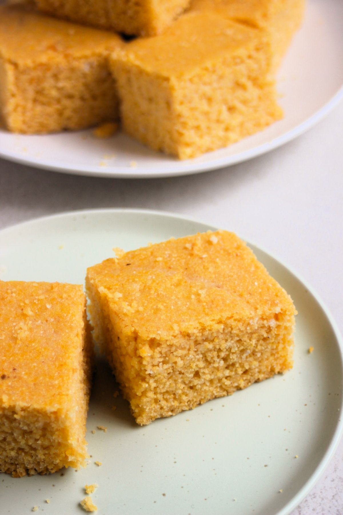 Two pieces of cornbread on an aqua-green plate. Behind, more cornbread pieces on a white plate.