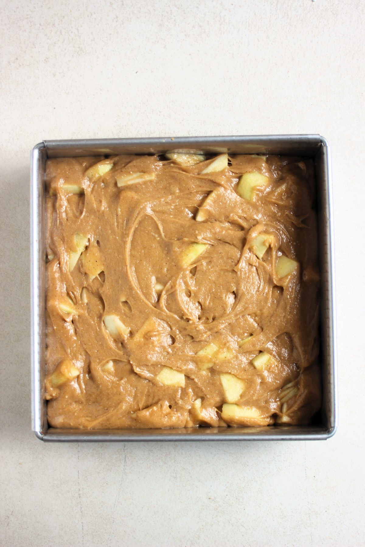 Square baking pan with a brown light batter that includes apples, before baking.