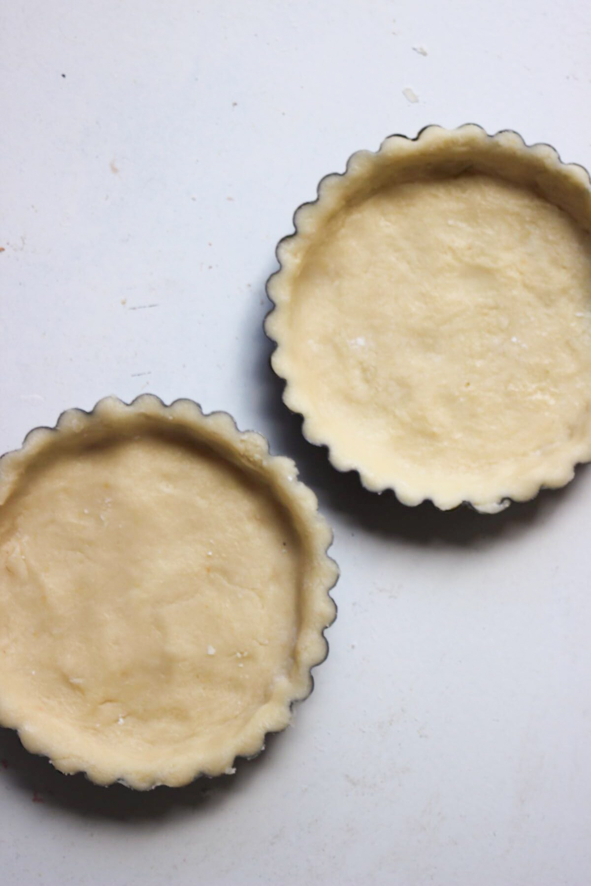 Two tartlet pans with raw crust dough inside on a white surface.