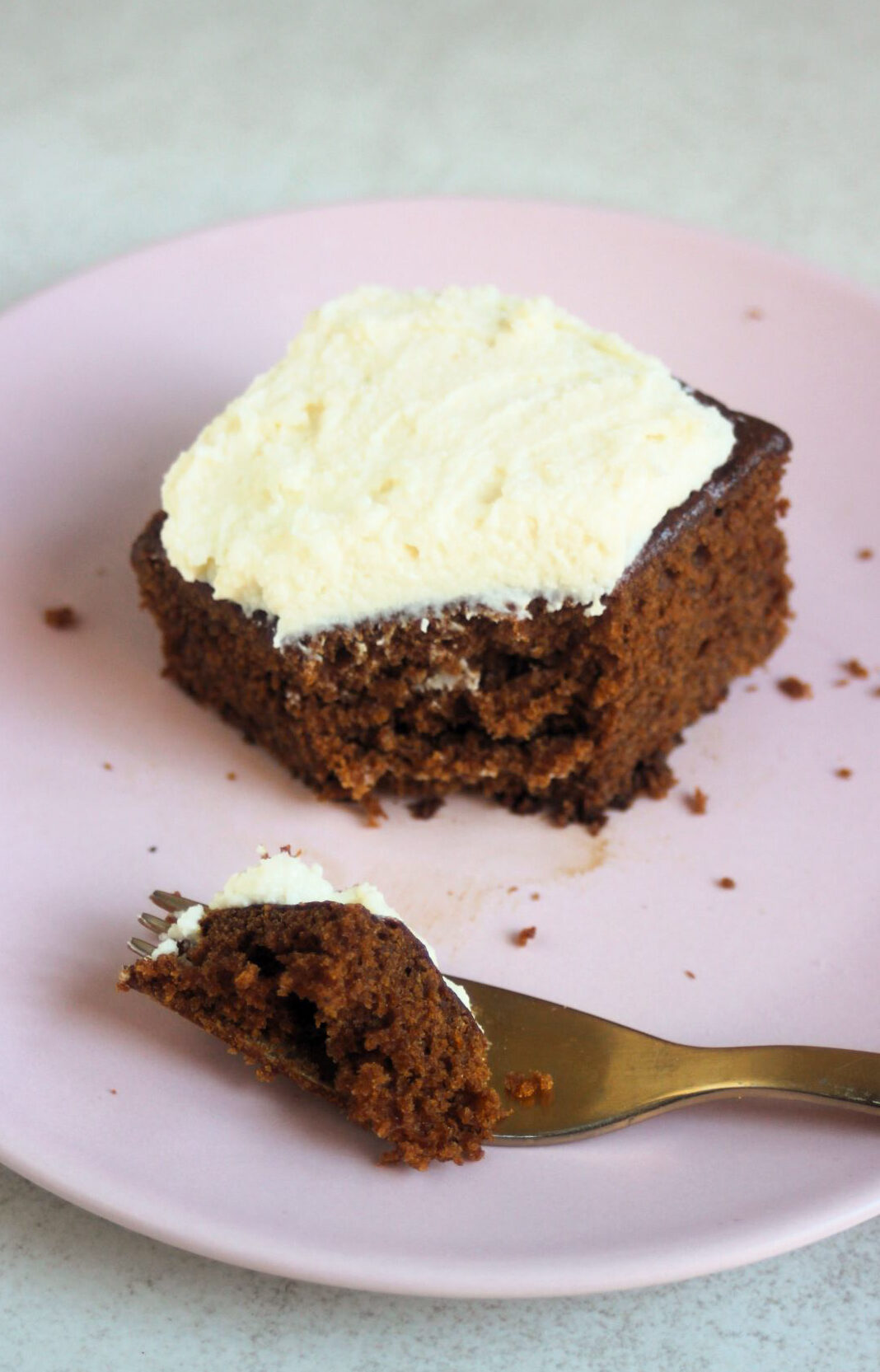 Portion of gingerbread cake with cream cheese frosting on a pink plate and golden fork with a piece of the cake.