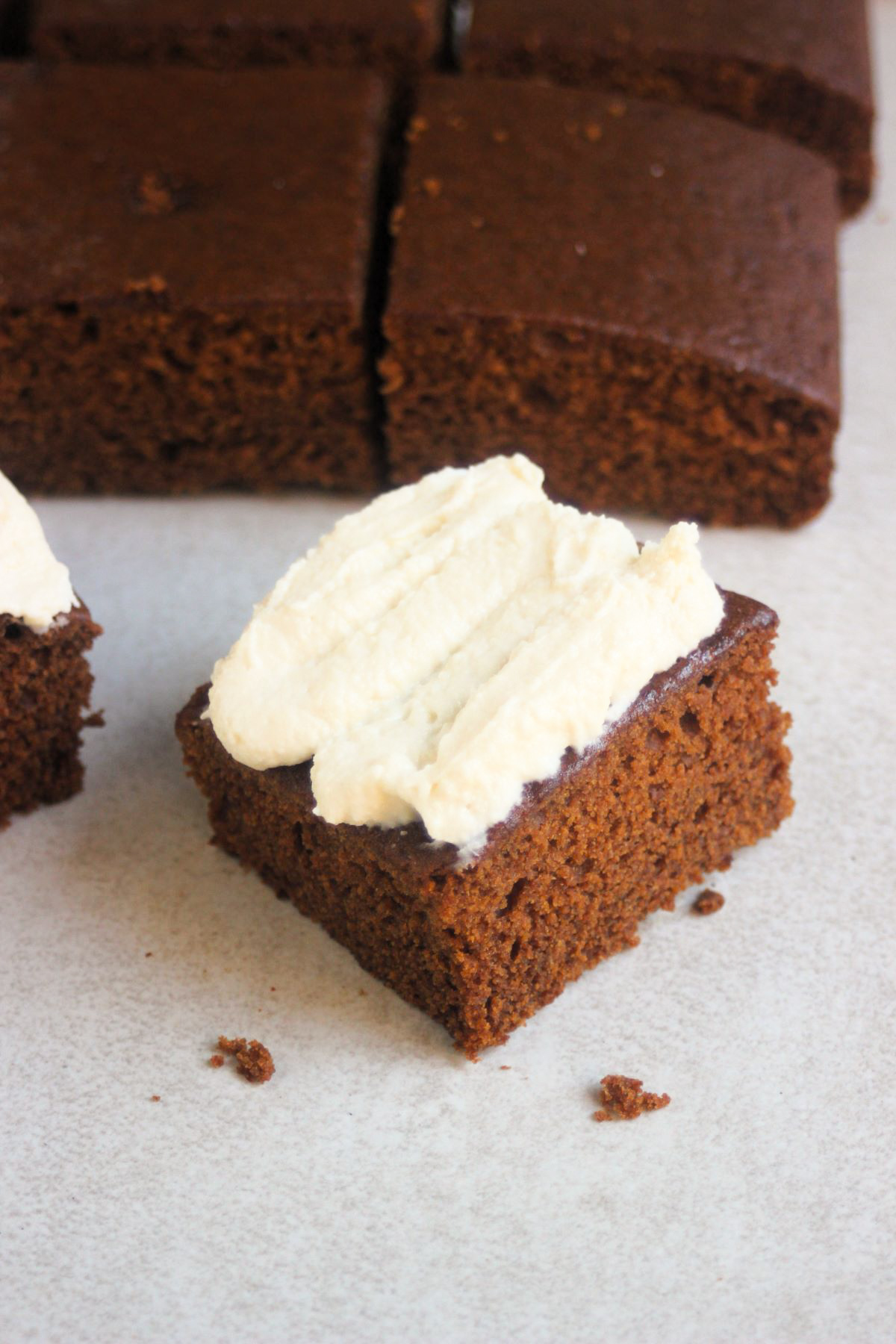 Portion of gingerbread cake with cream cheese frosting on a white surface. Gingerbread cake behind.