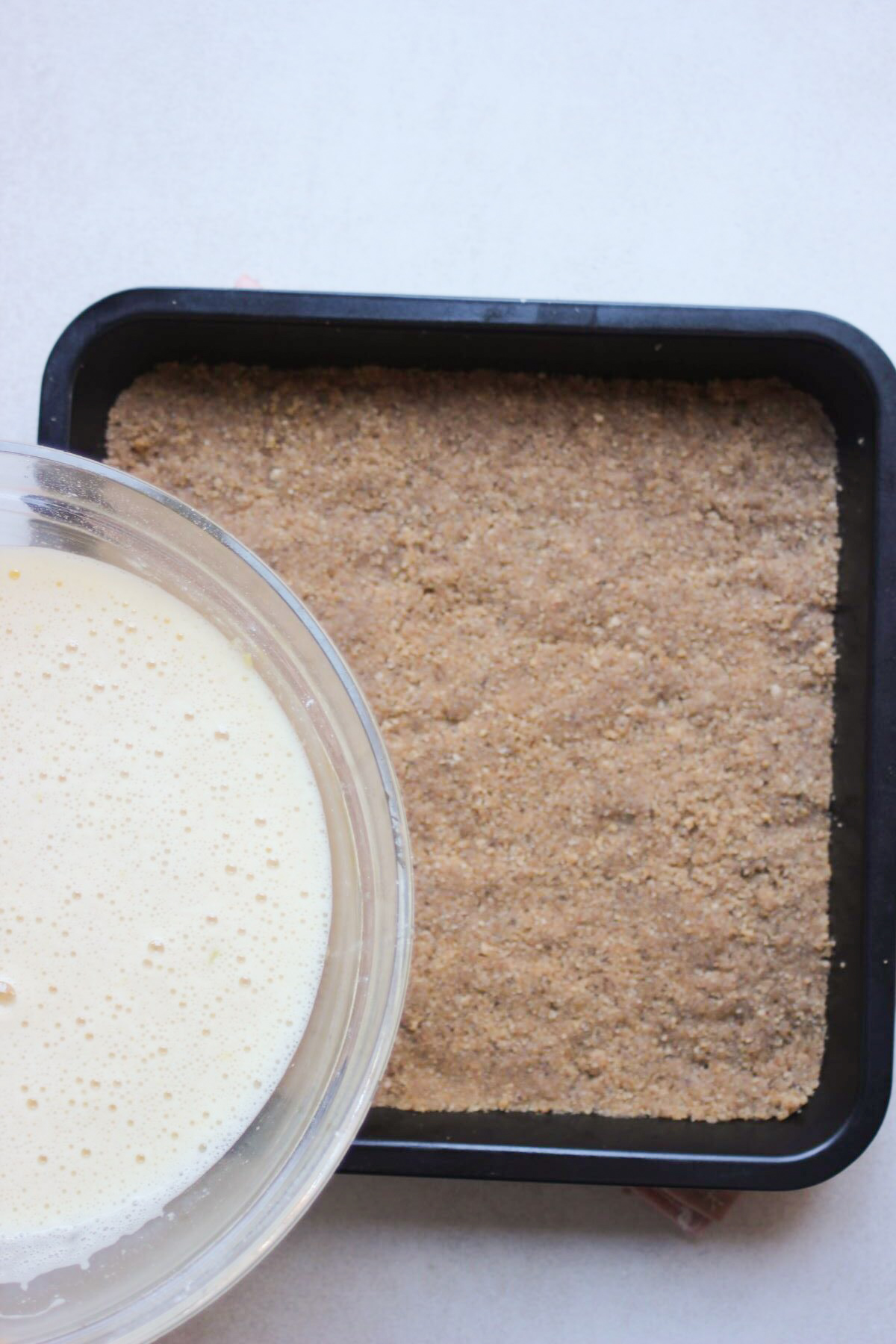 A glass bowl with liquid is about to be poured into a square baking pan with a cookie base.