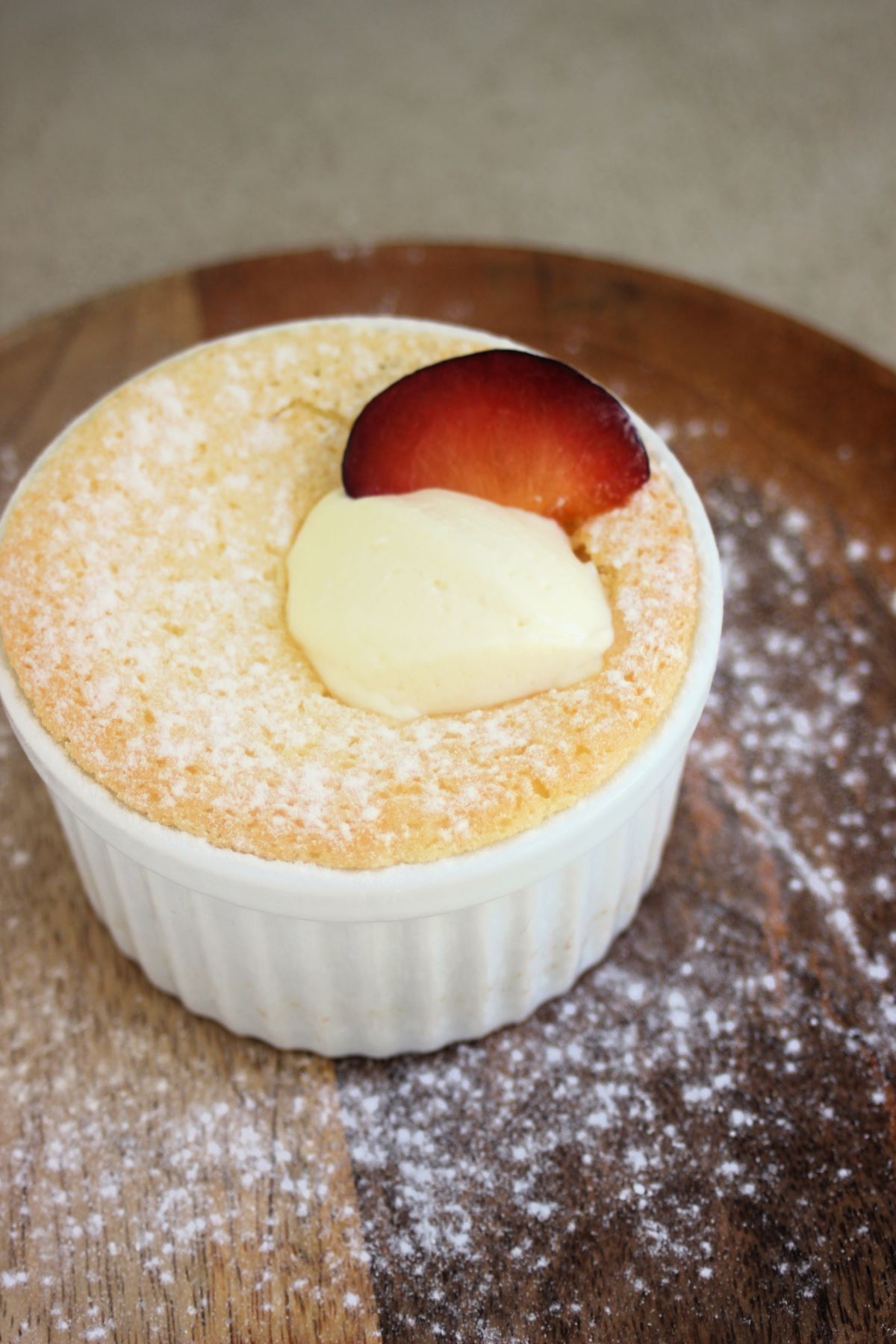 White ramekin with lemon pudding cake, whipped cream, and a plum slice on a wooden plate.