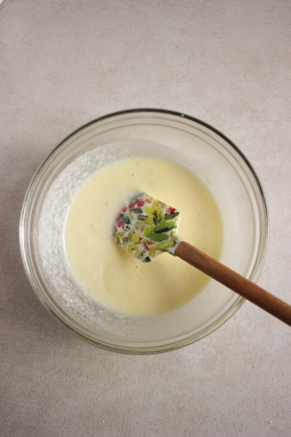 Glass bowl with a light yellow mixture and a rubber spatula on a white surface.
