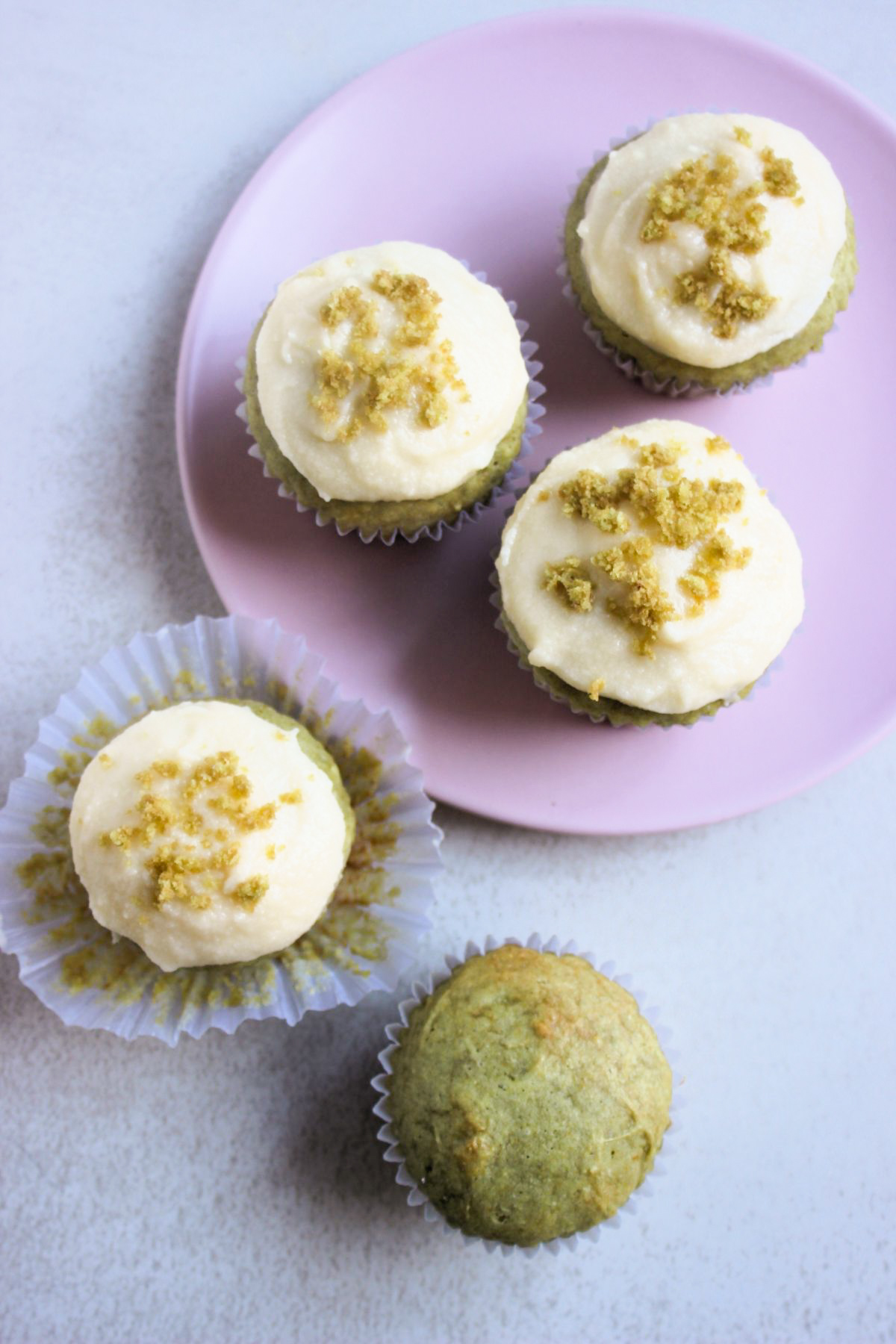A matcha cupcake without frosting and a matcha cupcake with the liner open, seen from above. On the side, more cupcakes on a pink plate.