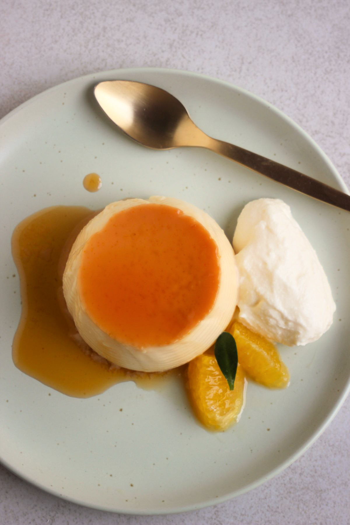 Orange flan with caramel, whipped cream, and fresh orange slices, on an aqua-green plate seen from above. A golden spoon beside.