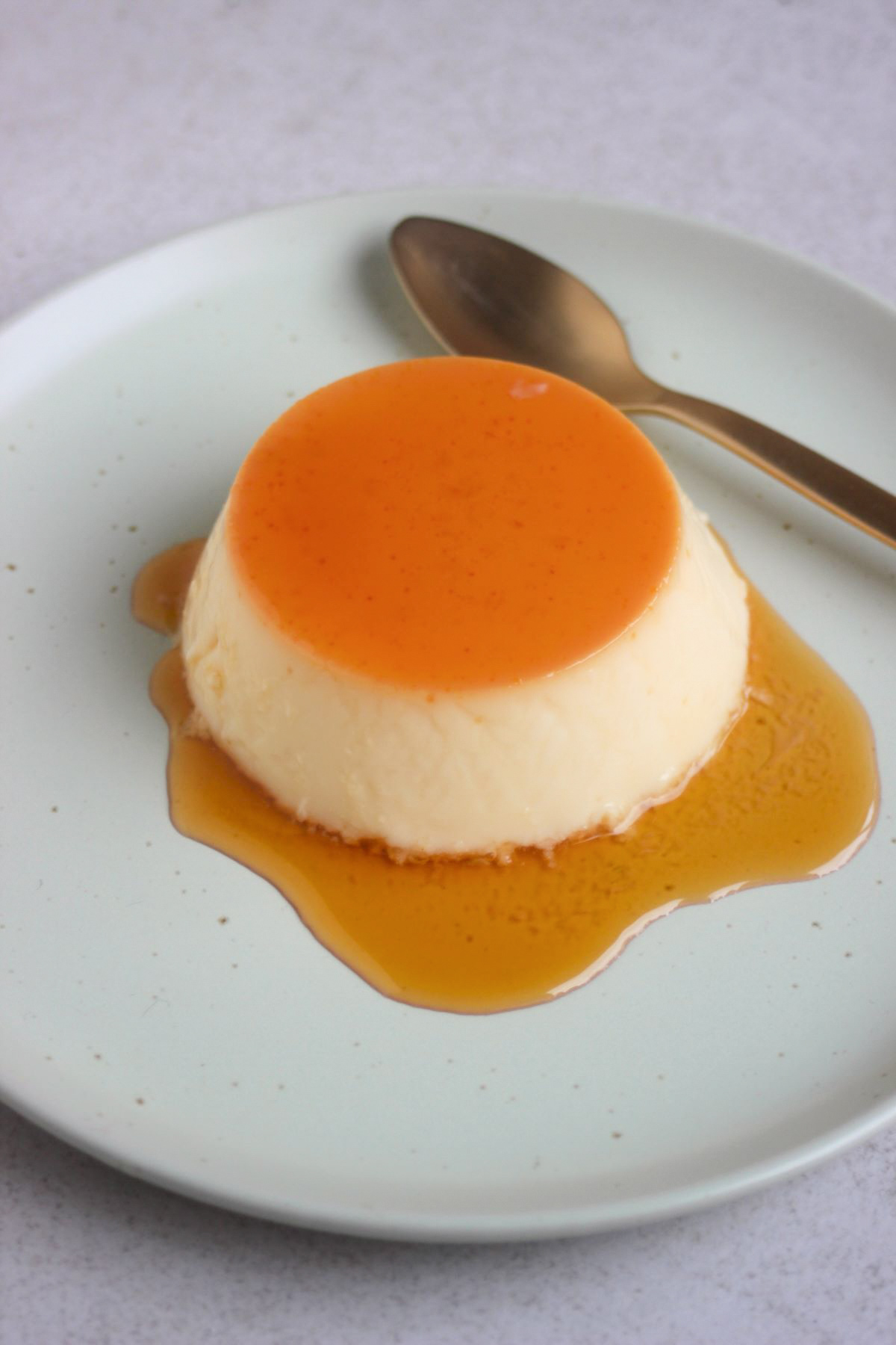 Orange flan with caramel on an aqua-green plate and a golden spoon.