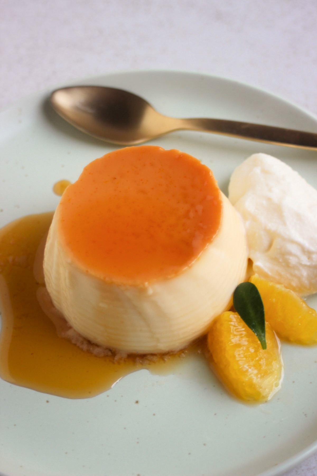 Orange flan with caramel, whipped cream, and fresh orange slices, on an aqua-green plate. A golden spoon behind.