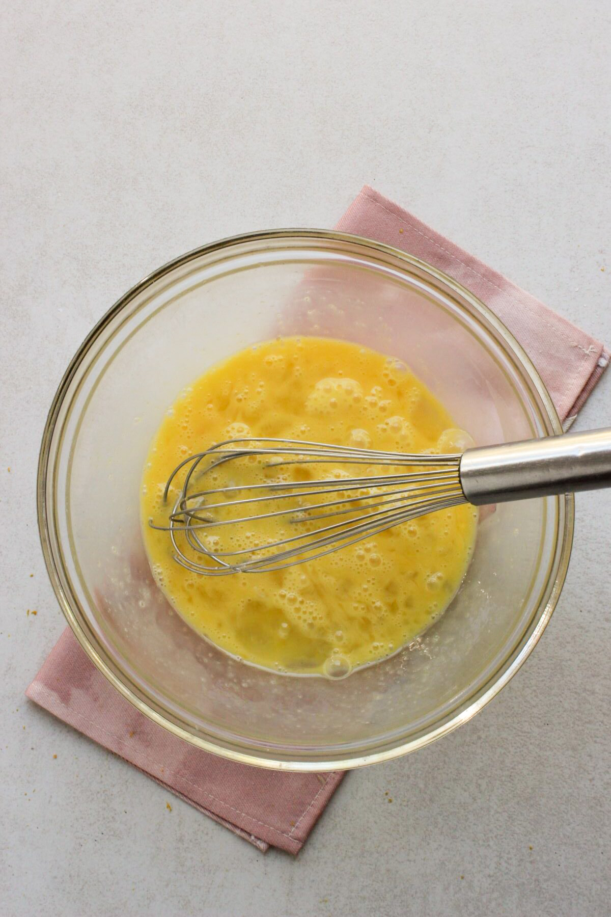 Glass bowl with eggs beaten and a hand whisk. Pink napkin under the bowl.
