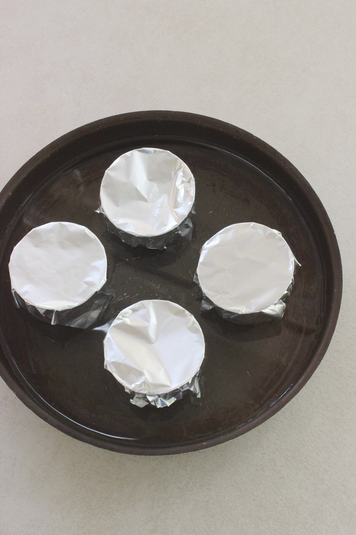 Four individual flan molds covered with aluminium on a baking sheet with water.