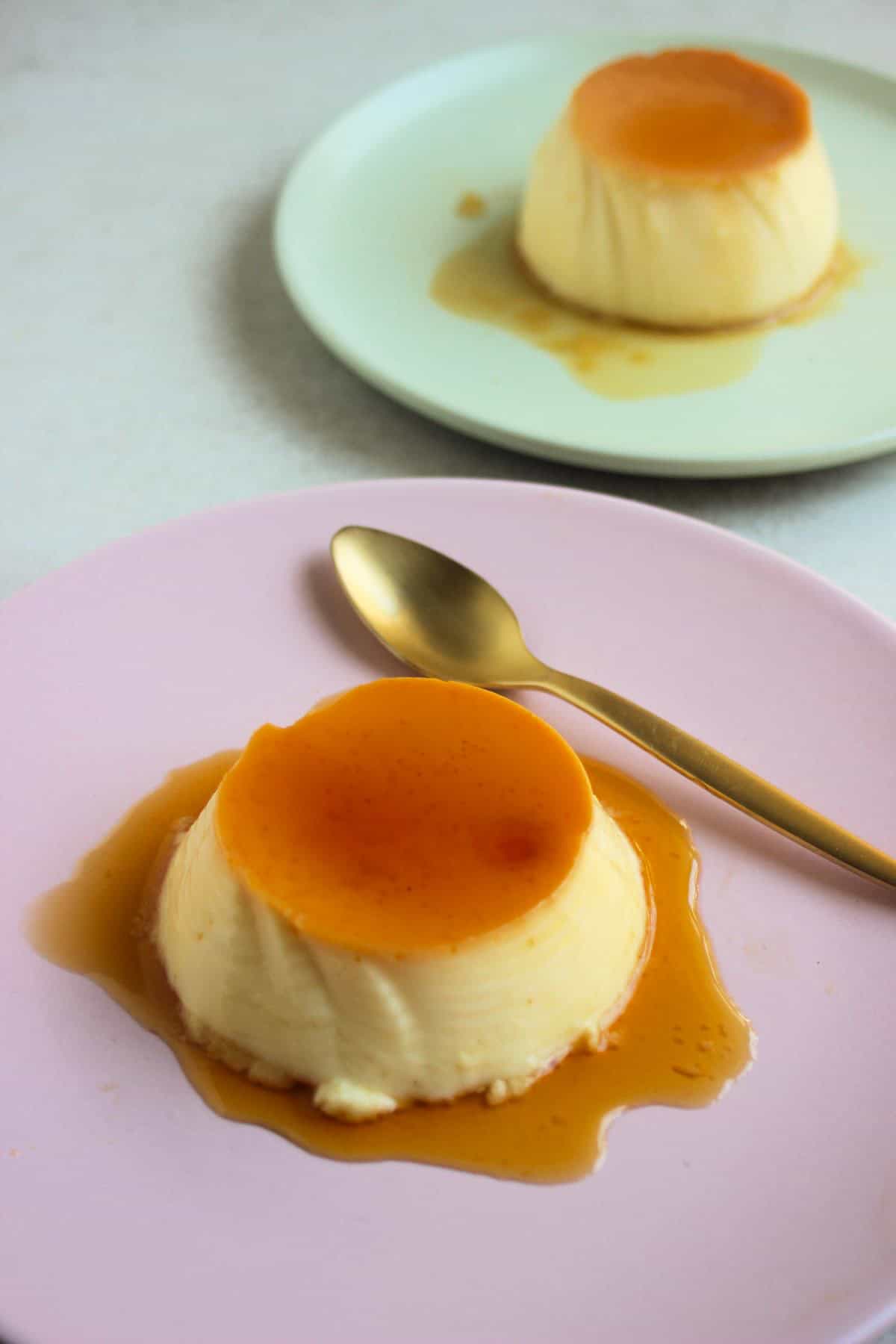 Orange flan with caramel on a pink plate and a golden spoon. Behind, another plate with flan with caramel.