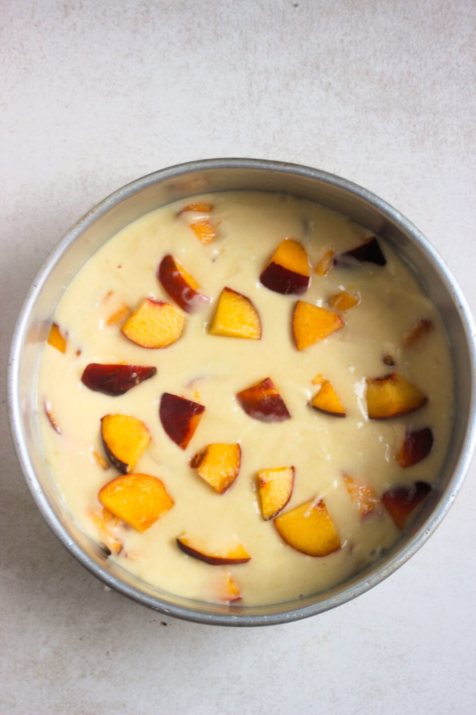 Round baking pan with cake batter and sliced peaches before baking seen from above.
