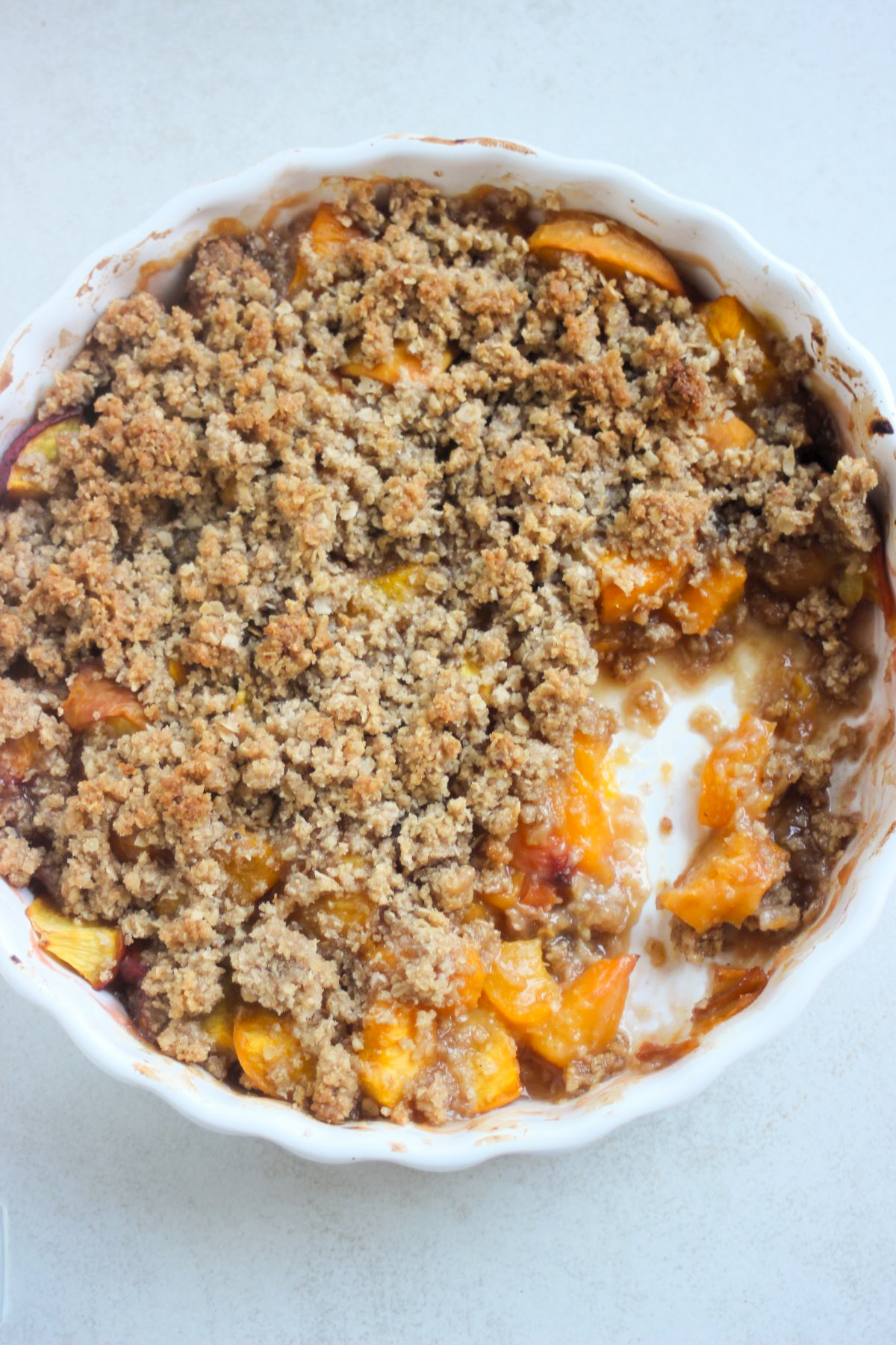 White round baking dish with peach crisp with a quarter eaten, on a white surface seen from above.