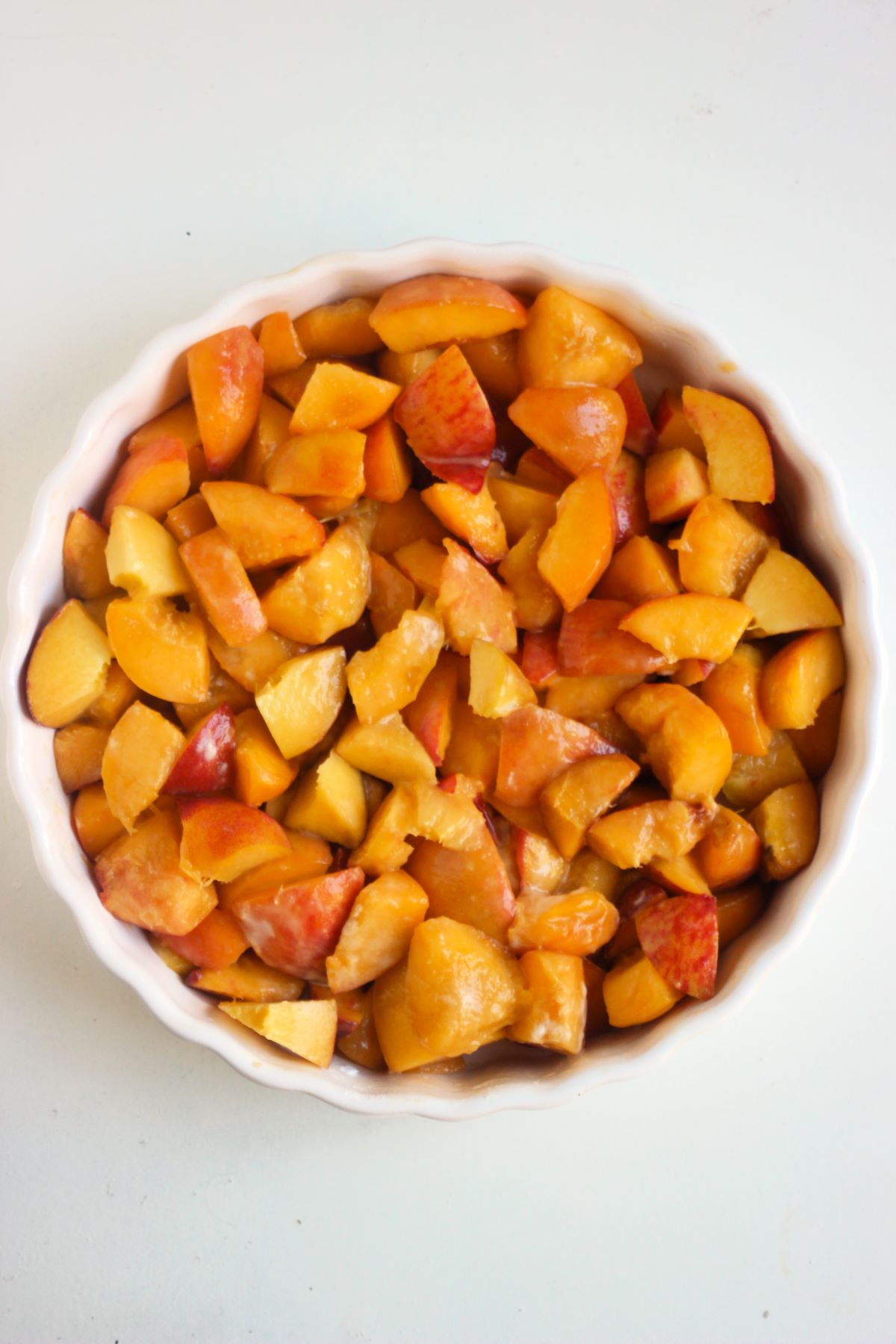 White round baking dish with sliced peaches on a white surface seen form above.