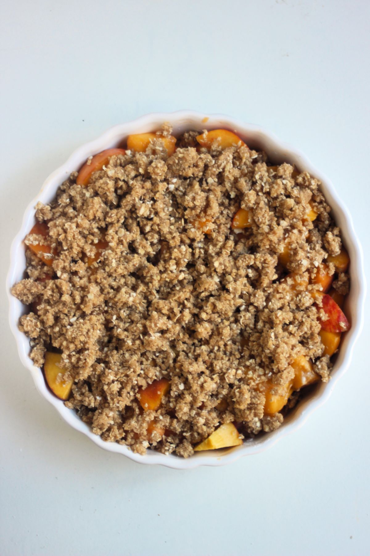 White round baking dish with raw peach crisp on a white surface seen from above.