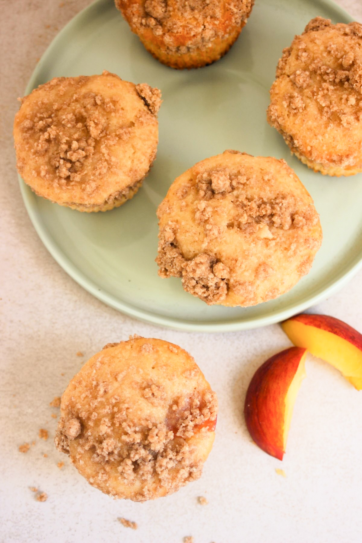 Peach streusel muffins on an aqua-green plate seen from above. A muffin and fresh peach slices on the side.