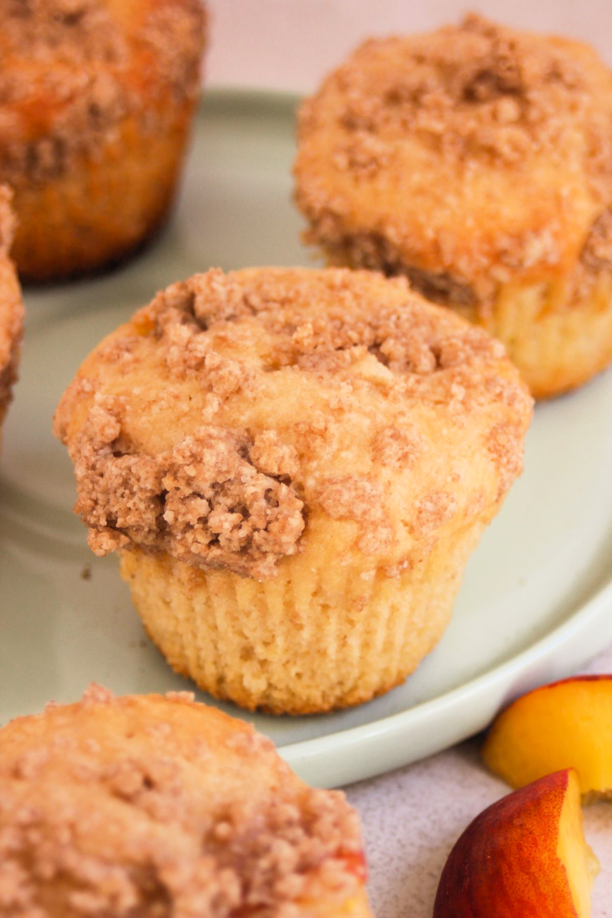Peach streusel muffin on an aqua green plate, More muffins, and fresh peach slices on the sides.