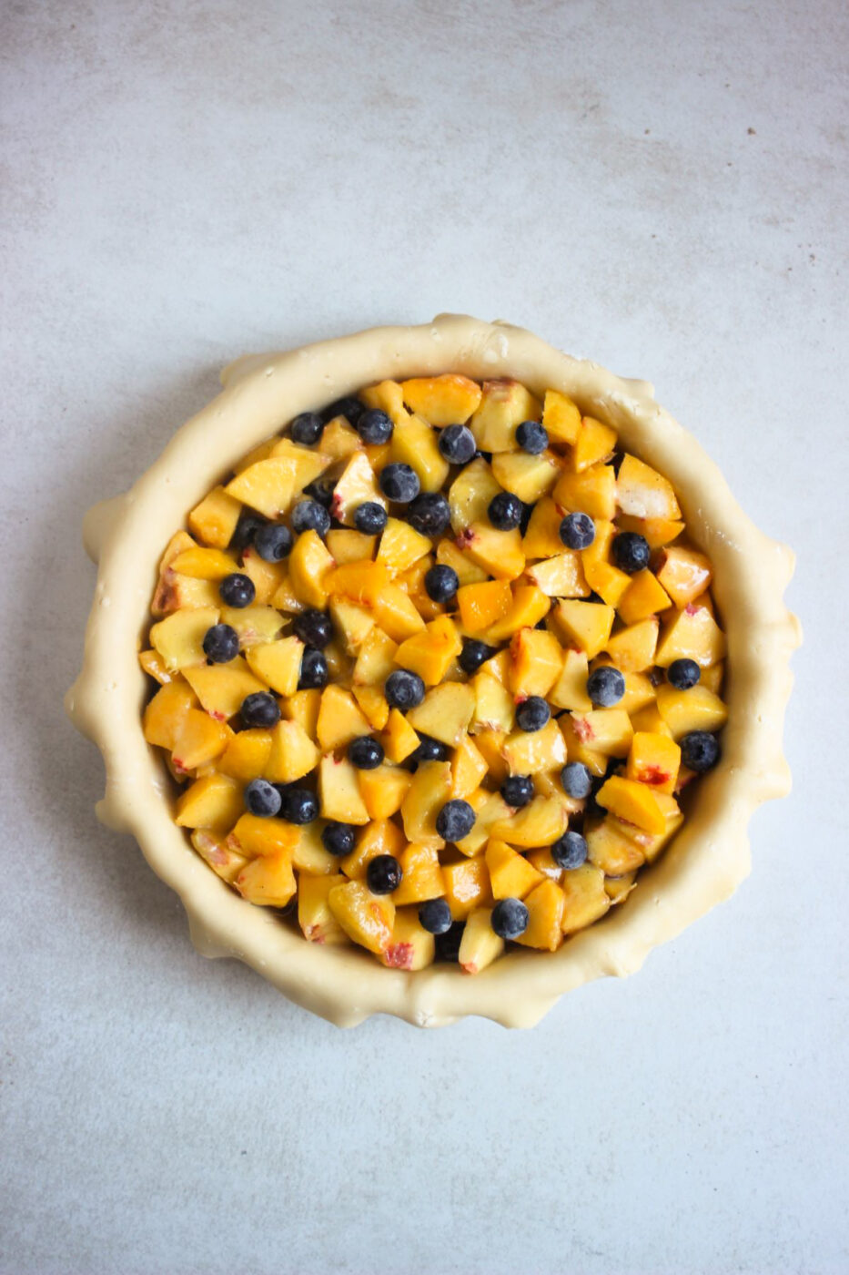 Peach pie raw batter on a round baking dish with peach slices and blueberries seen from above.